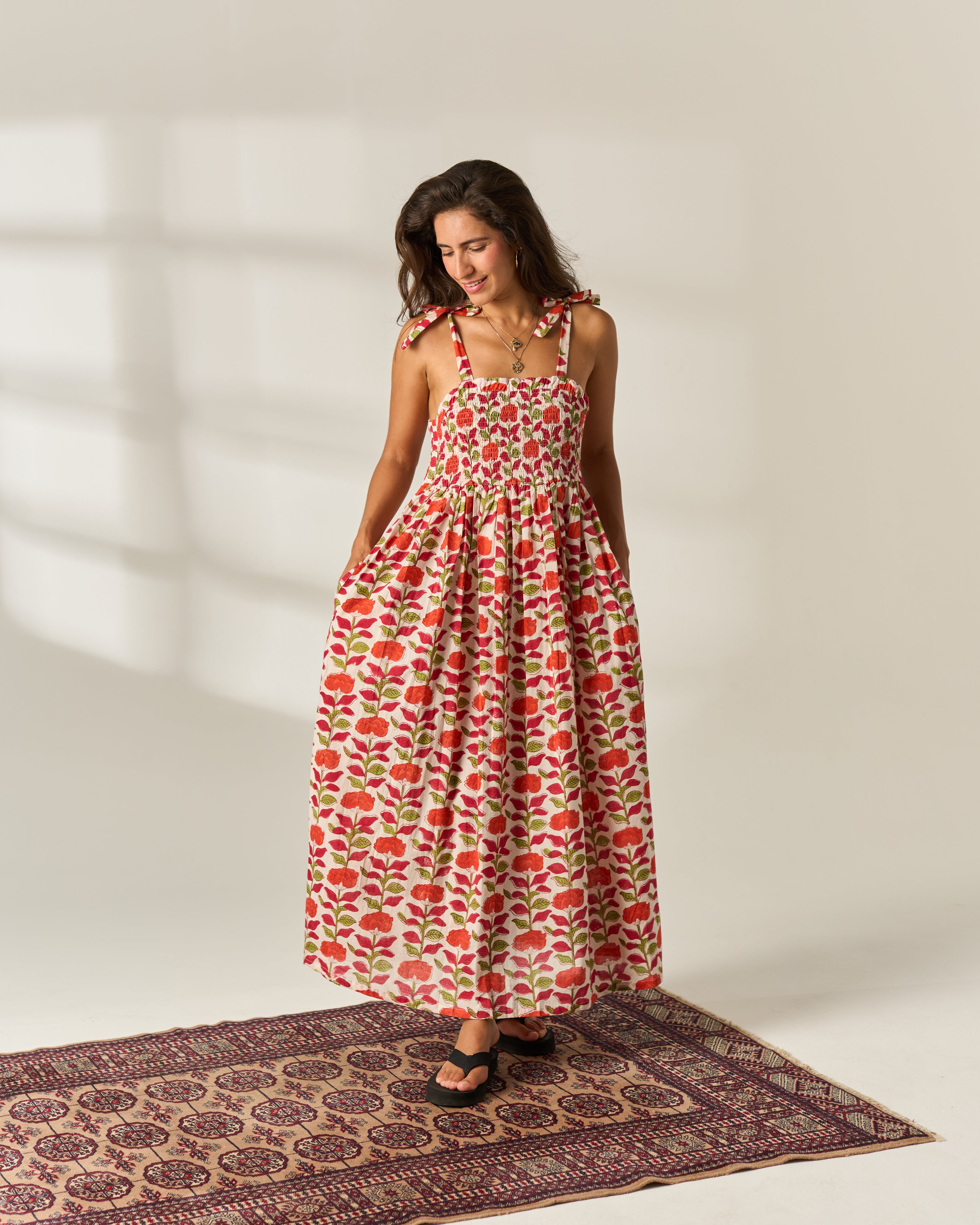 A woman wearing a shoulder strapped, smocked maxi dress with a red and green floral hand block print on a cream cotton fabric base, standing on a patterned rug against a warm off-white wall.