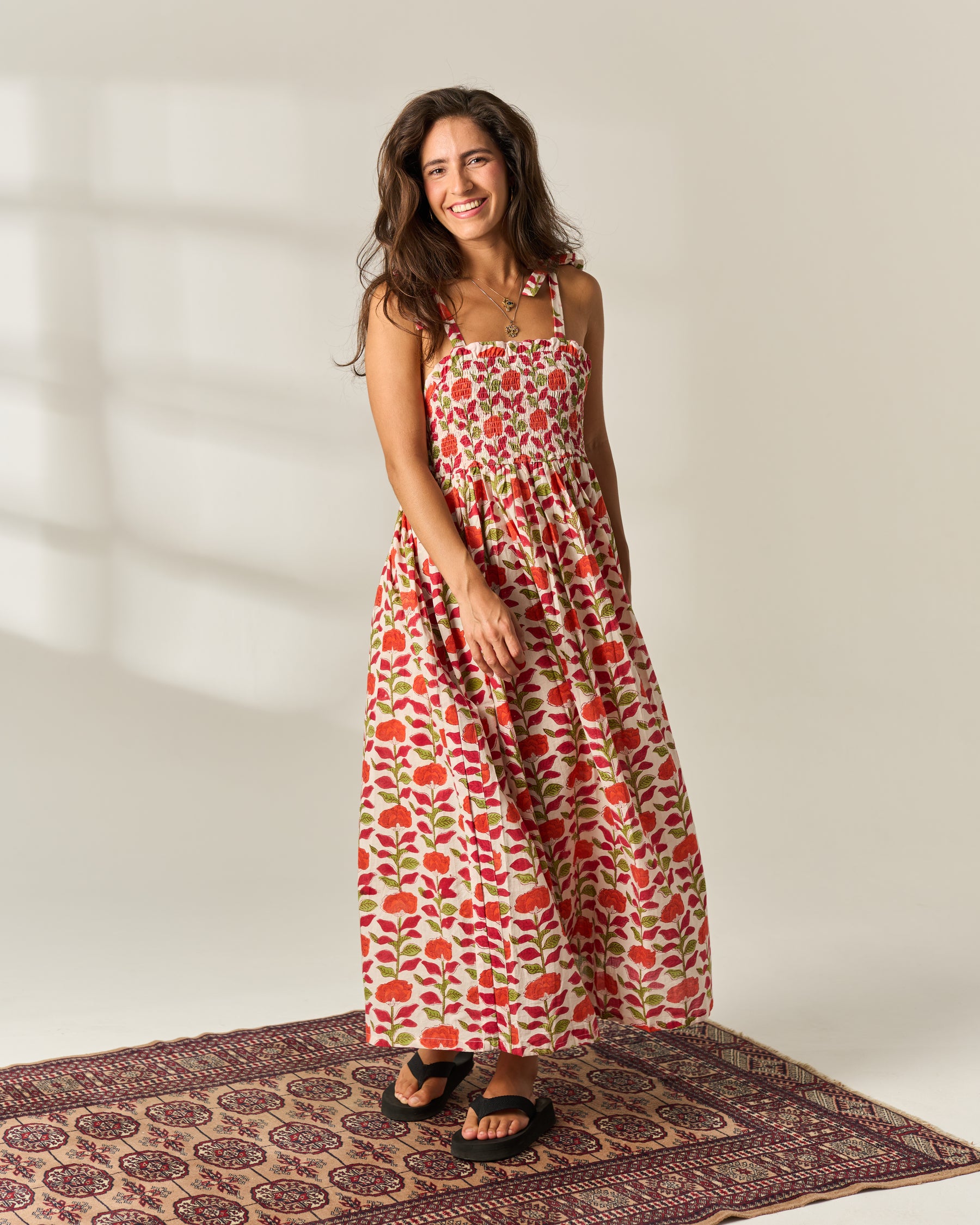 A woman modeling a shoulder strapped, smocked maxi dress with a red and green leaf pattern on a cream cotton fabric base, standing on a patterned rug.