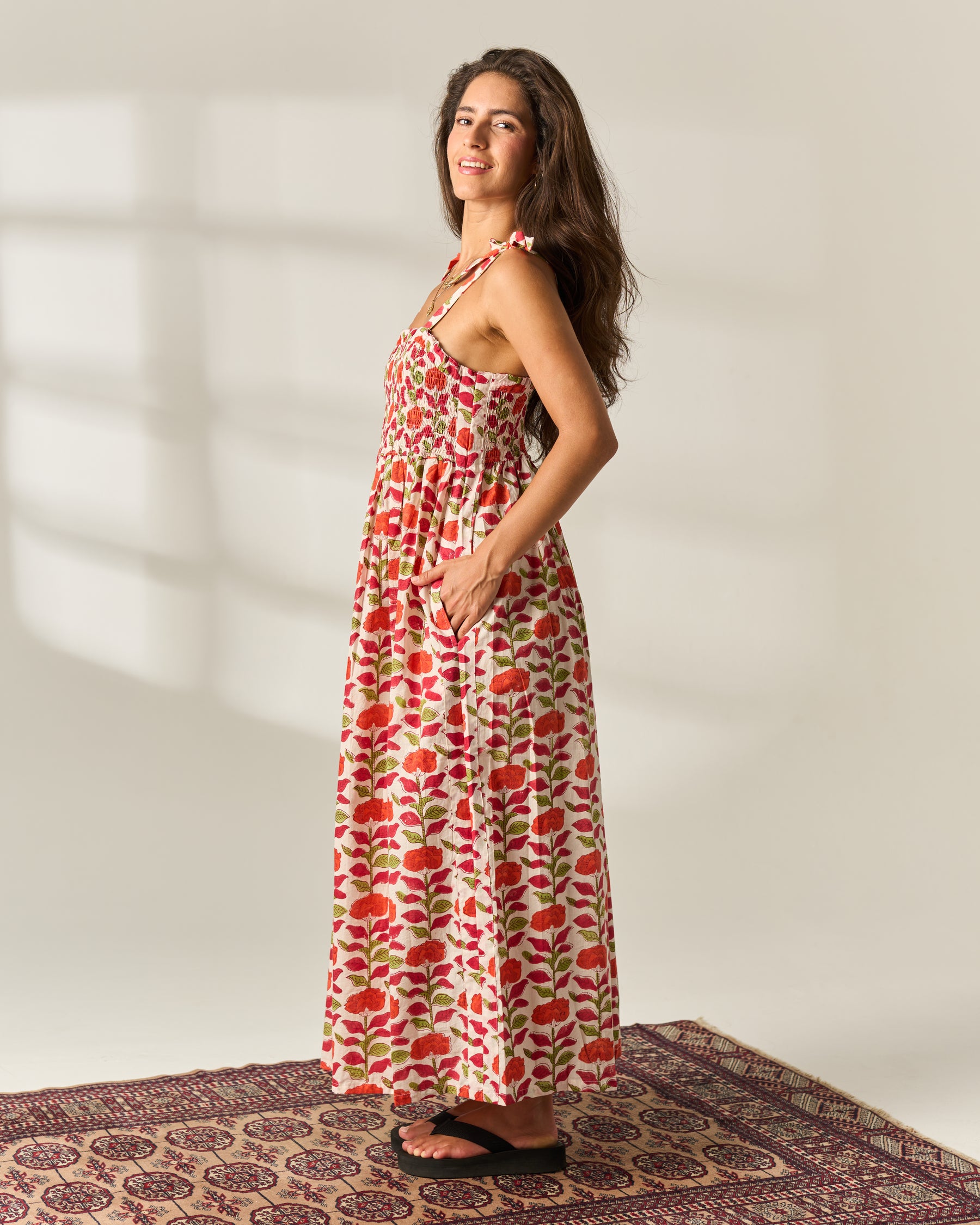 A woman modeling a sleeveless, strappy, floor-length maxi Hand Block Print dress with a red and green floral print on a cream cotton base fabric, standing on a patterned rug against a neutral wall with soft window shadows.