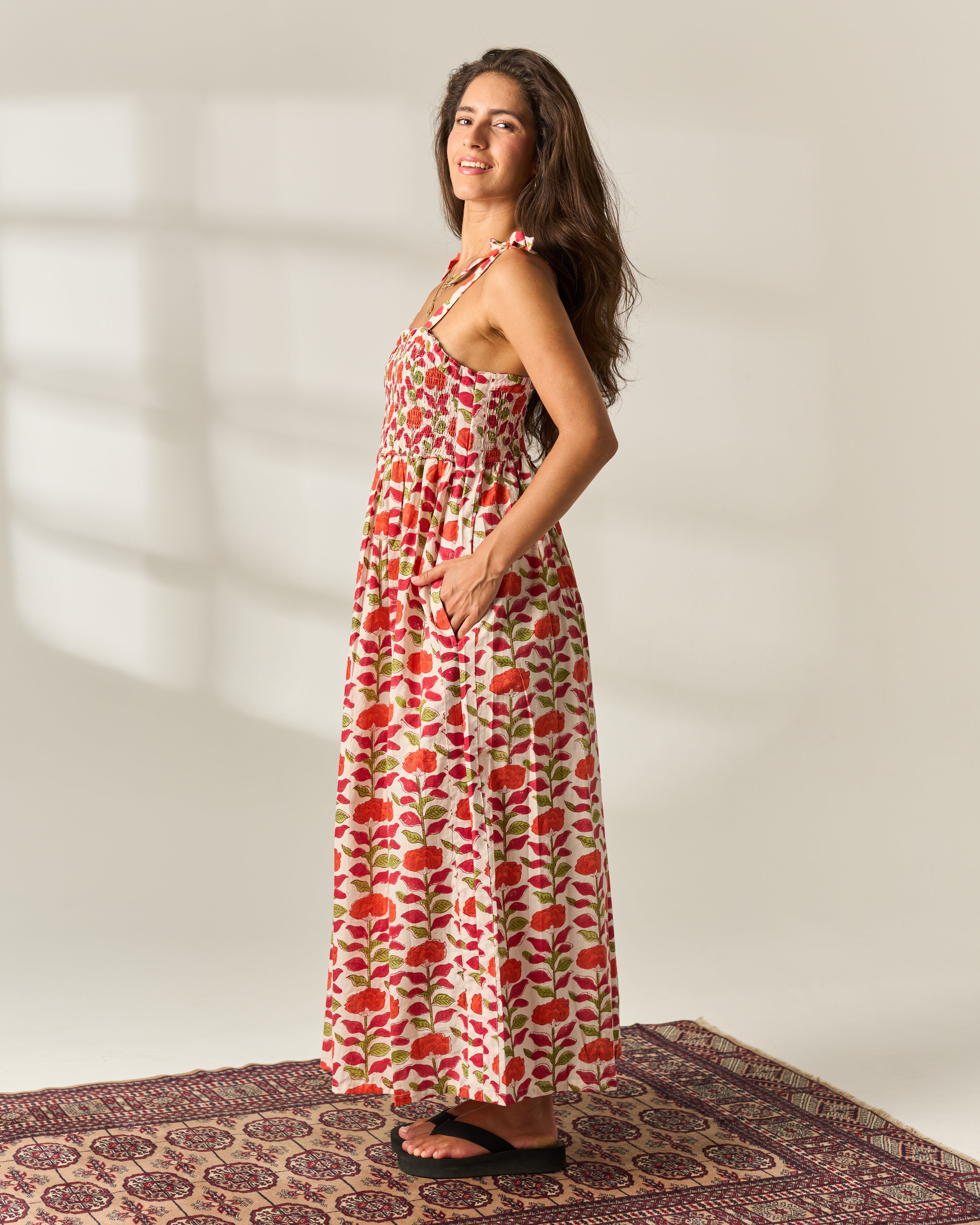 A woman modeling a sleeveless, strappy, floor-length maxi Hand Block Print dress with a red and green floral print on a cream cotton base fabric, standing on a patterned rug against a neutral wall with soft window shadows.