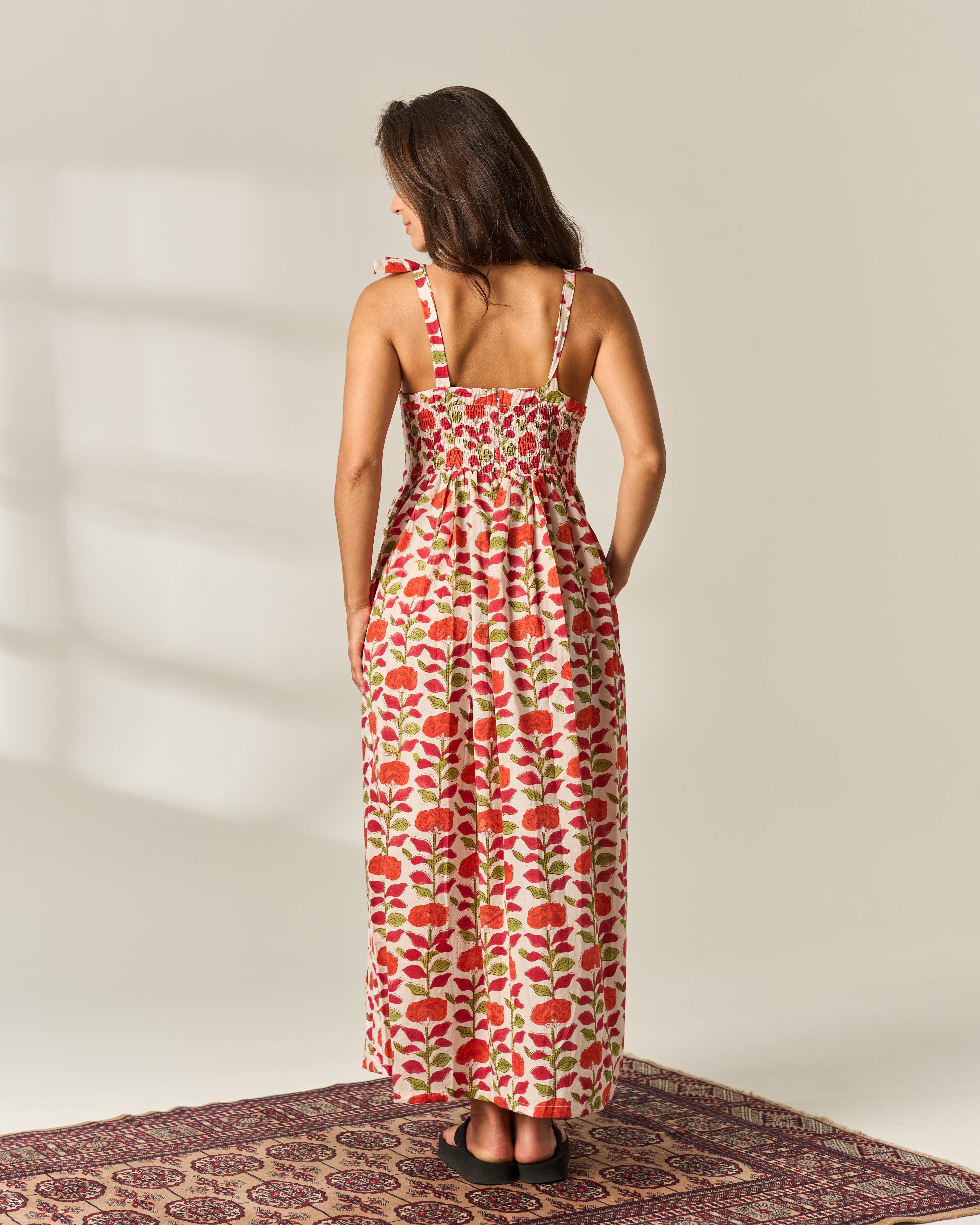 A woman stands with her back to the camera wearing a sleeveless strappy maxi dress with a red and green floral hand block painted pattern on a cream cotton fabric base. She is standing on a patterned rug against a plain beige wall with soft window shadows.