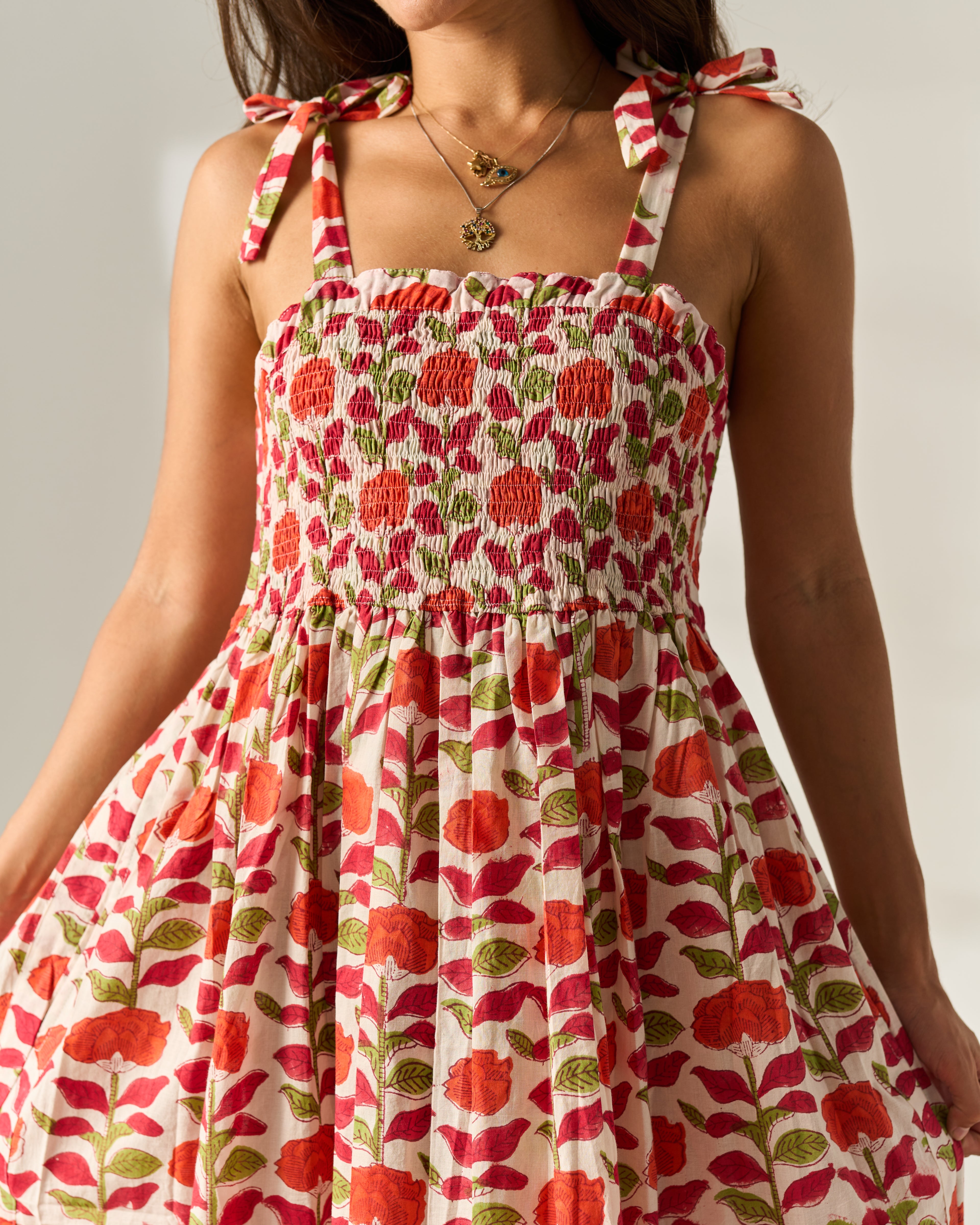 A close-up of a woman wearing a sleeveless shoulder strapped smocked maxi dress with tie straps, featuring a red and orange floral hand block print on a cream cotton fabric base.