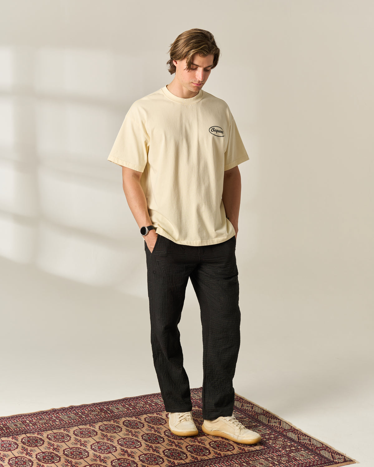 A man wearing a cream-colored short-sleeve t-shirt with a small graphic on the left chest, black pants, and cream sneakers, standing on a patterned rug against a neutral studio background.