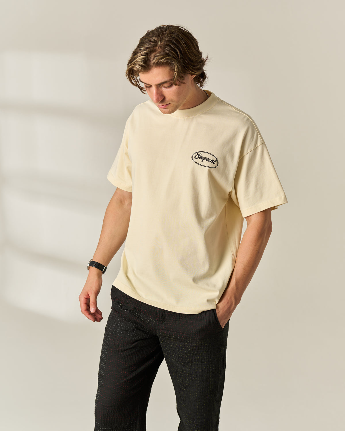 A man wearing a cream-colored short-sleeve t-shirt with a small black oval 'Sequent' logo on the left chest, paired with black pants, standing against a light beige background.