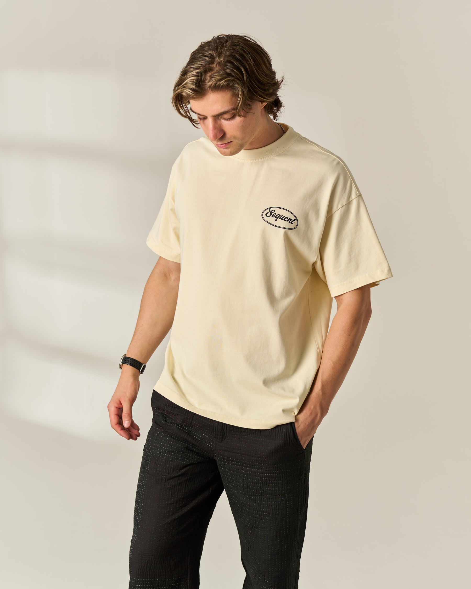 A man wearing a cream-colored short-sleeve t-shirt with a small black oval 'Sequent' logo on the left chest, paired with black pants, standing against a light beige background.