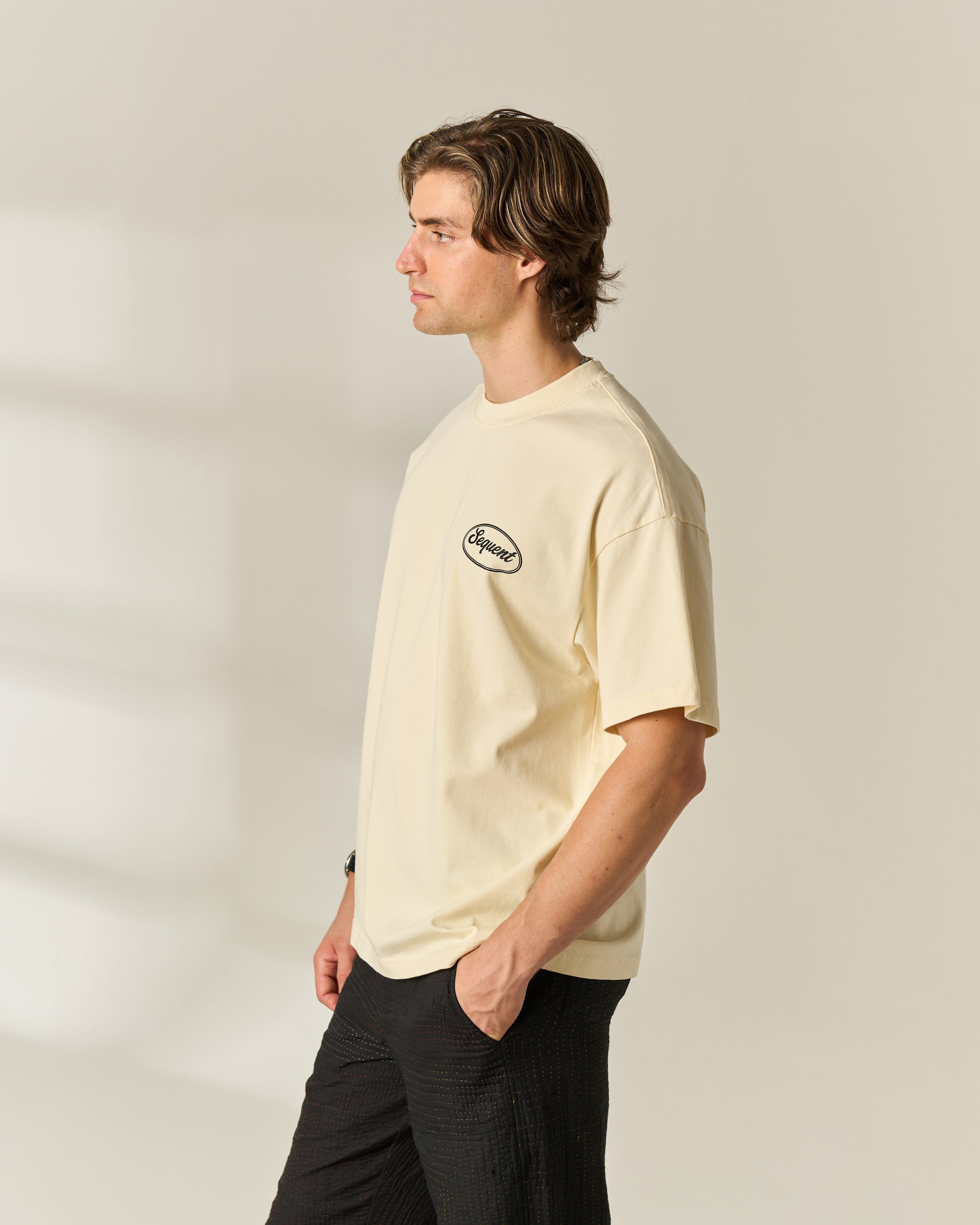 A man in profile wearing a cream-colored short-sleeve t-shirt with a small black oval chest 'Sequent' logo, paired with black pants, standing against a light beige studio background.