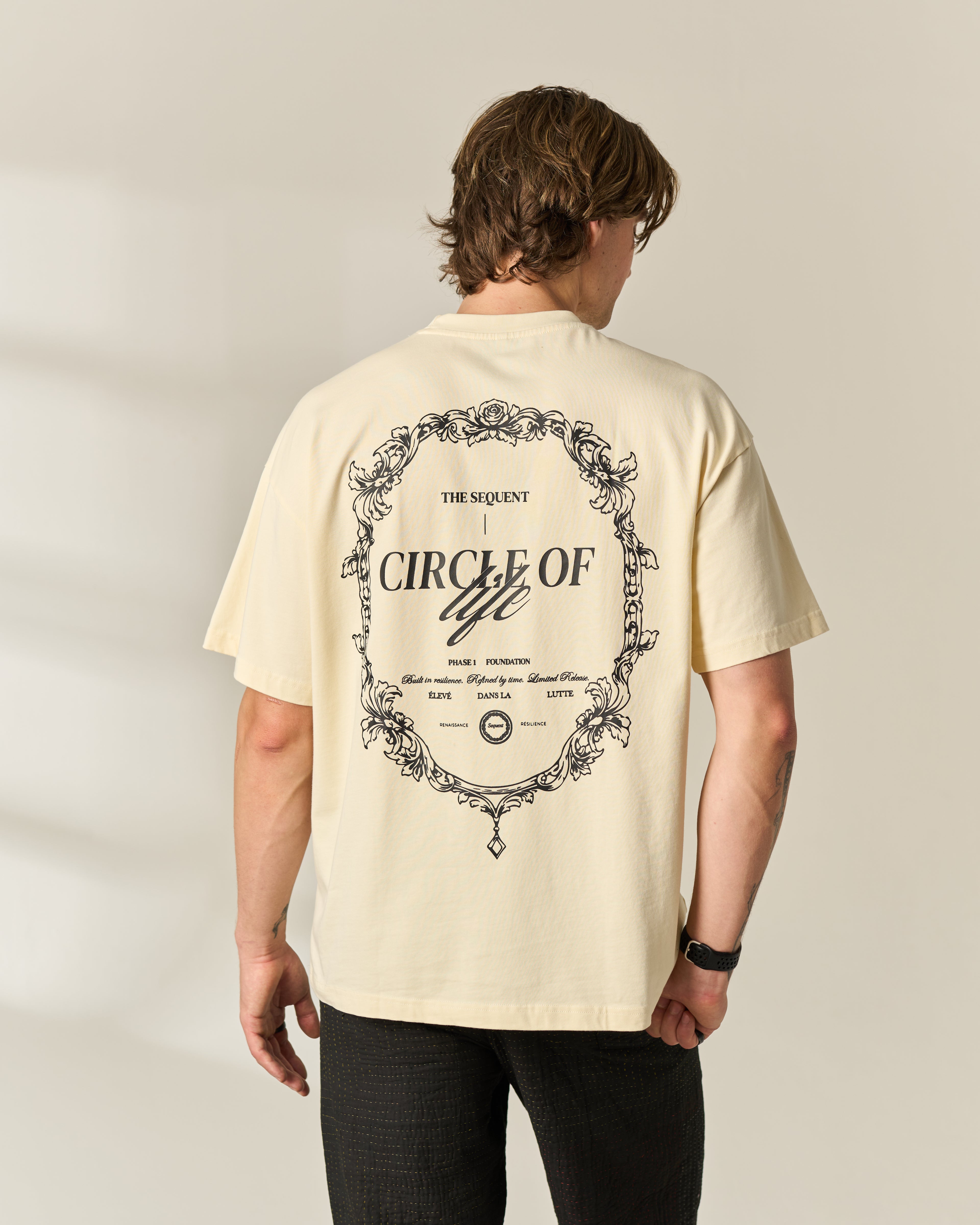 A man viewed from behind wearing a cream-colored t-shirt with a large black graphic print on the back about 'Circle of Life', paired with black pants, against a neutral beige background.