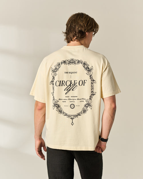 A man viewed from behind wearing a cream-colored t-shirt with a large black graphic print on the back about 'Circle of Life', paired with black pants, against a neutral beige background.