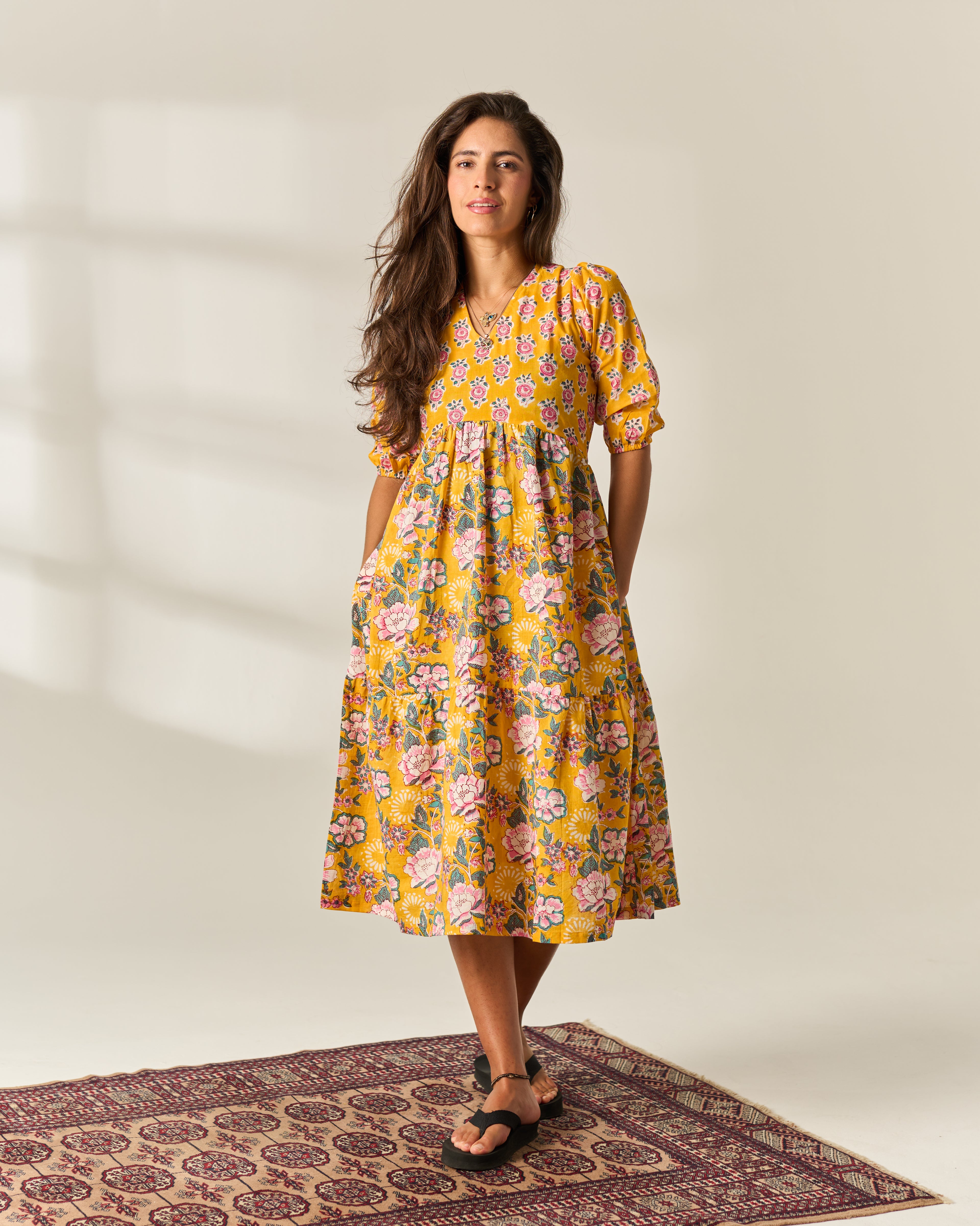 A woman wearing a yellow midi hand block painted dress with a pink and green floral print, standing on a patterned rug against a warm off-white wall.