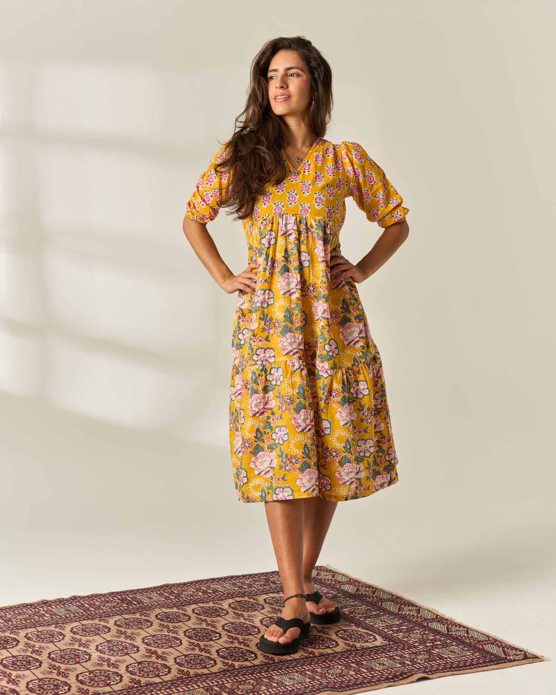 a woman modeling for a mustard and floral hand block printed midi dress, posing on a textured rug against a white wall