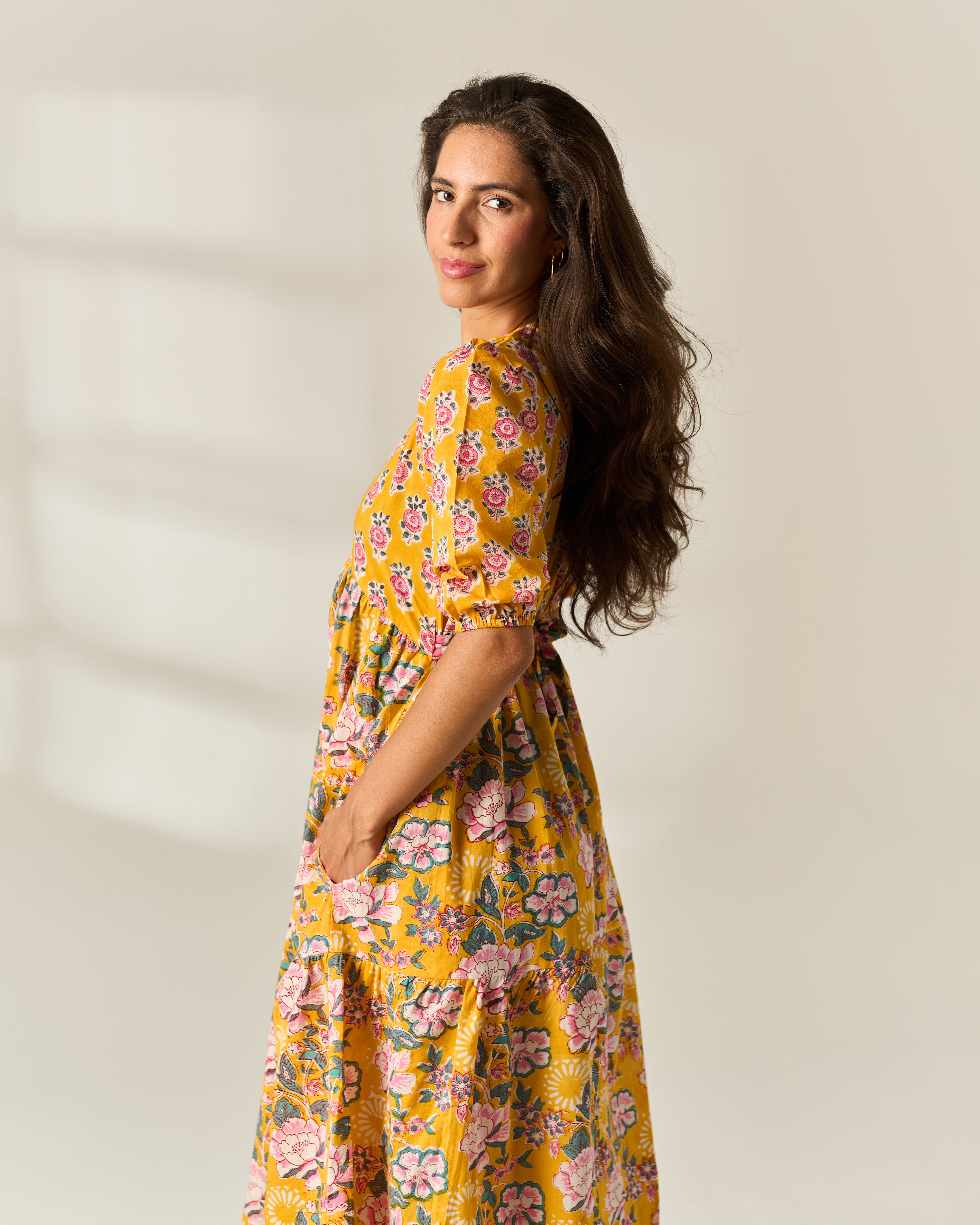 A woman with long dark wavy hair is shown from the side/back wearing a yellow floral midi hand block painted dress with pink and green flowers, standing against a plain light beige wall.