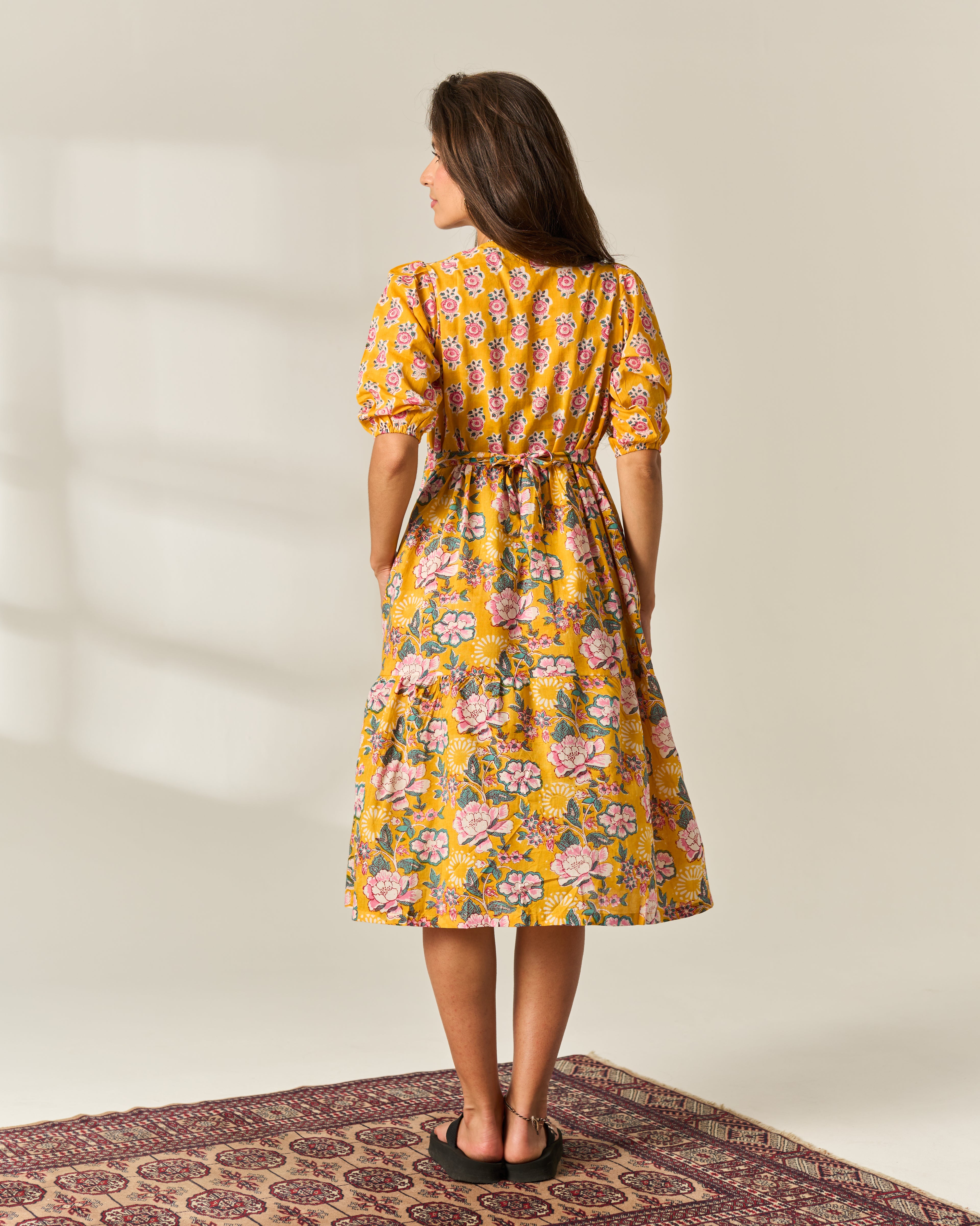 A woman stands with her back to the camera wearing a mustard yellow midi dress with a pink and green floral hand block painted pattern, short puff sleeves, and a tie waist, set against a neutral studio background with a patterned rug.