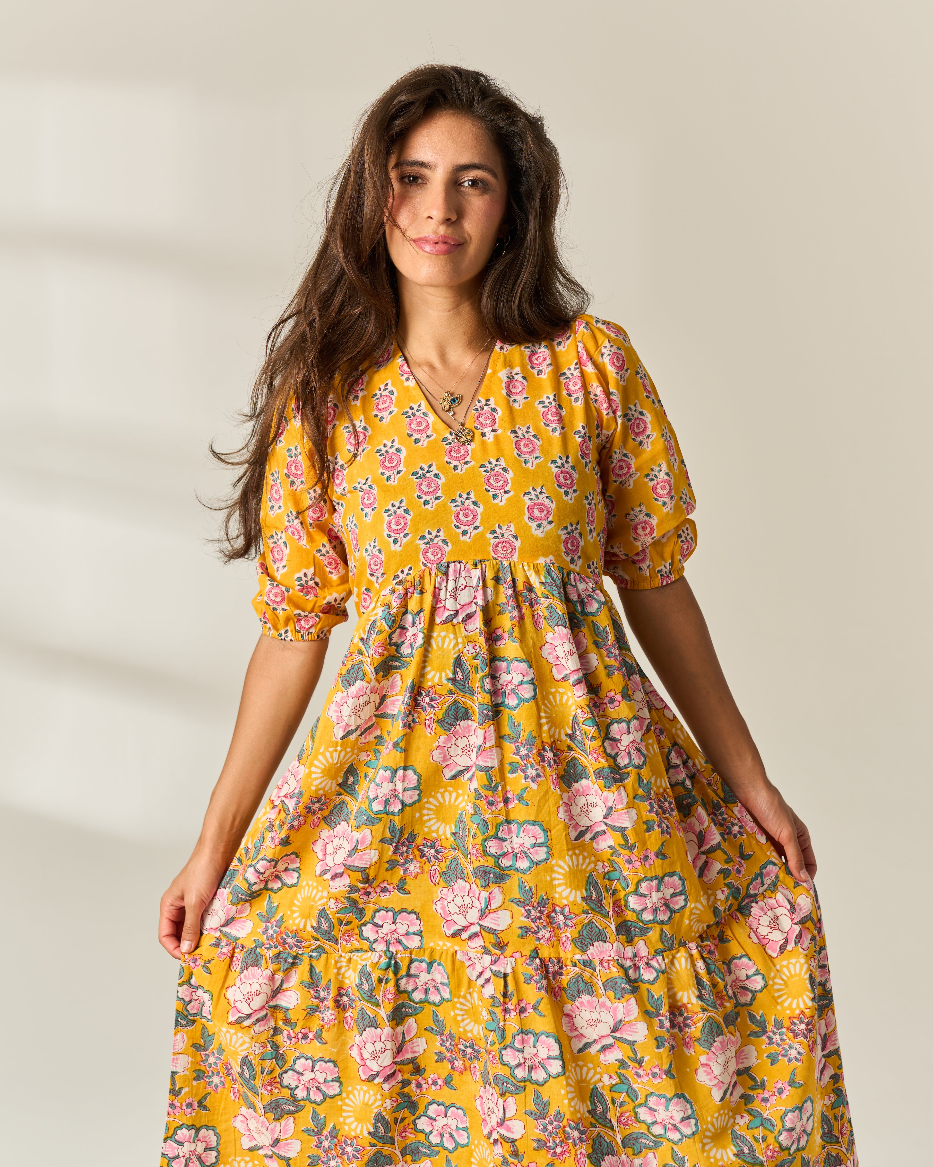A woman wearing a yellow tiered midi dress with a pink and green floral print, standing against a light beige background.
