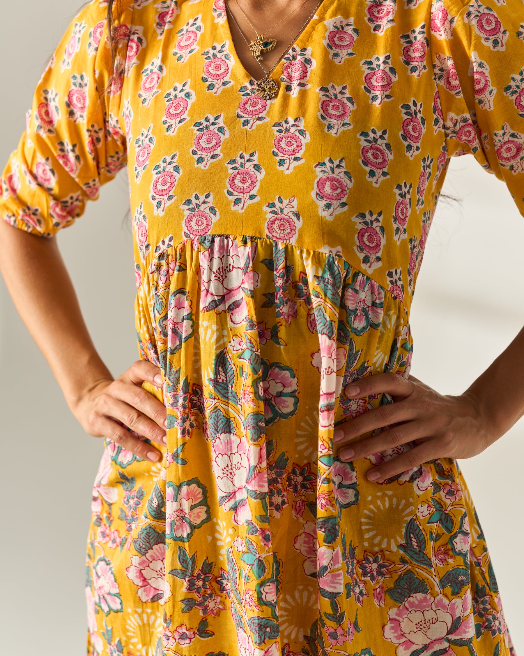 A close-up of a woman wearing a yellow floral dress with pink and green flowers, standing with hands on hips against a light neutral background.