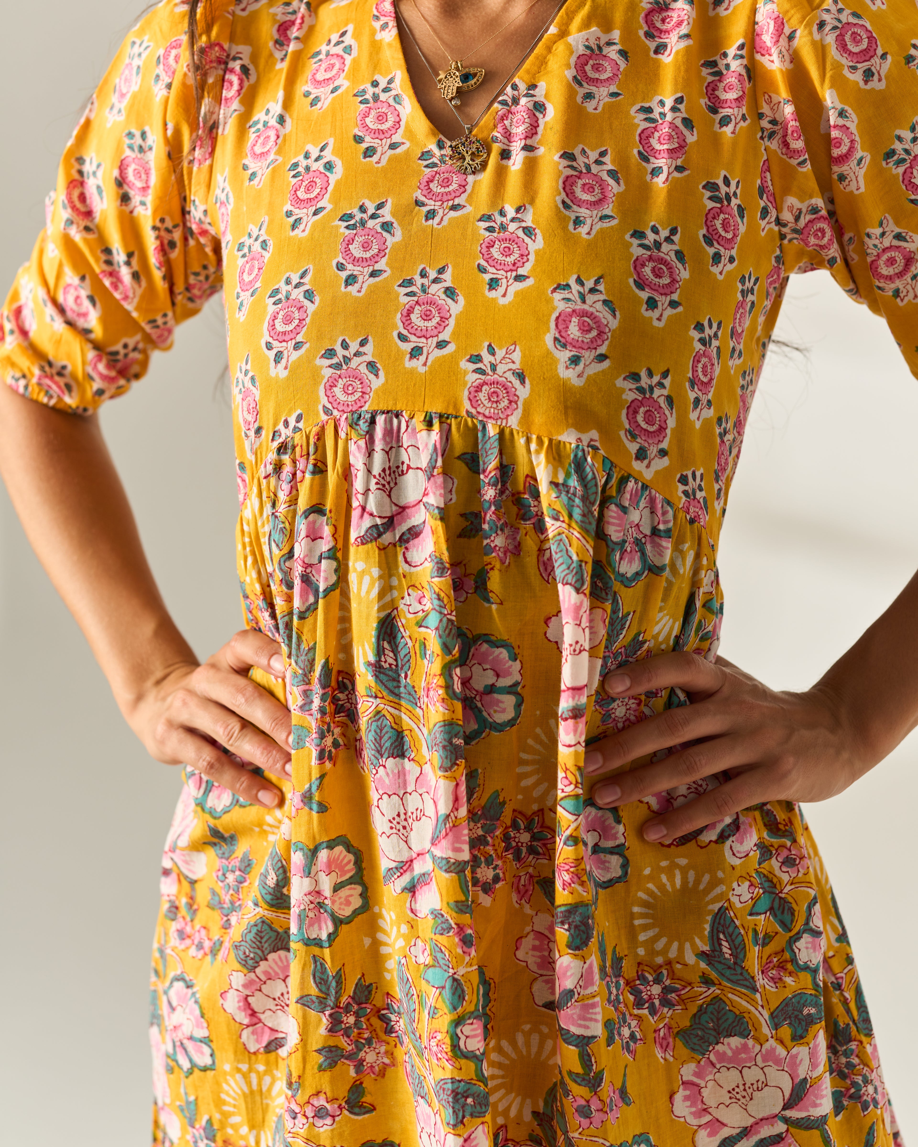 A close-up of a woman wearing a yellow floral dress with pink and green flowers, standing with hands on hips against a light neutral background.