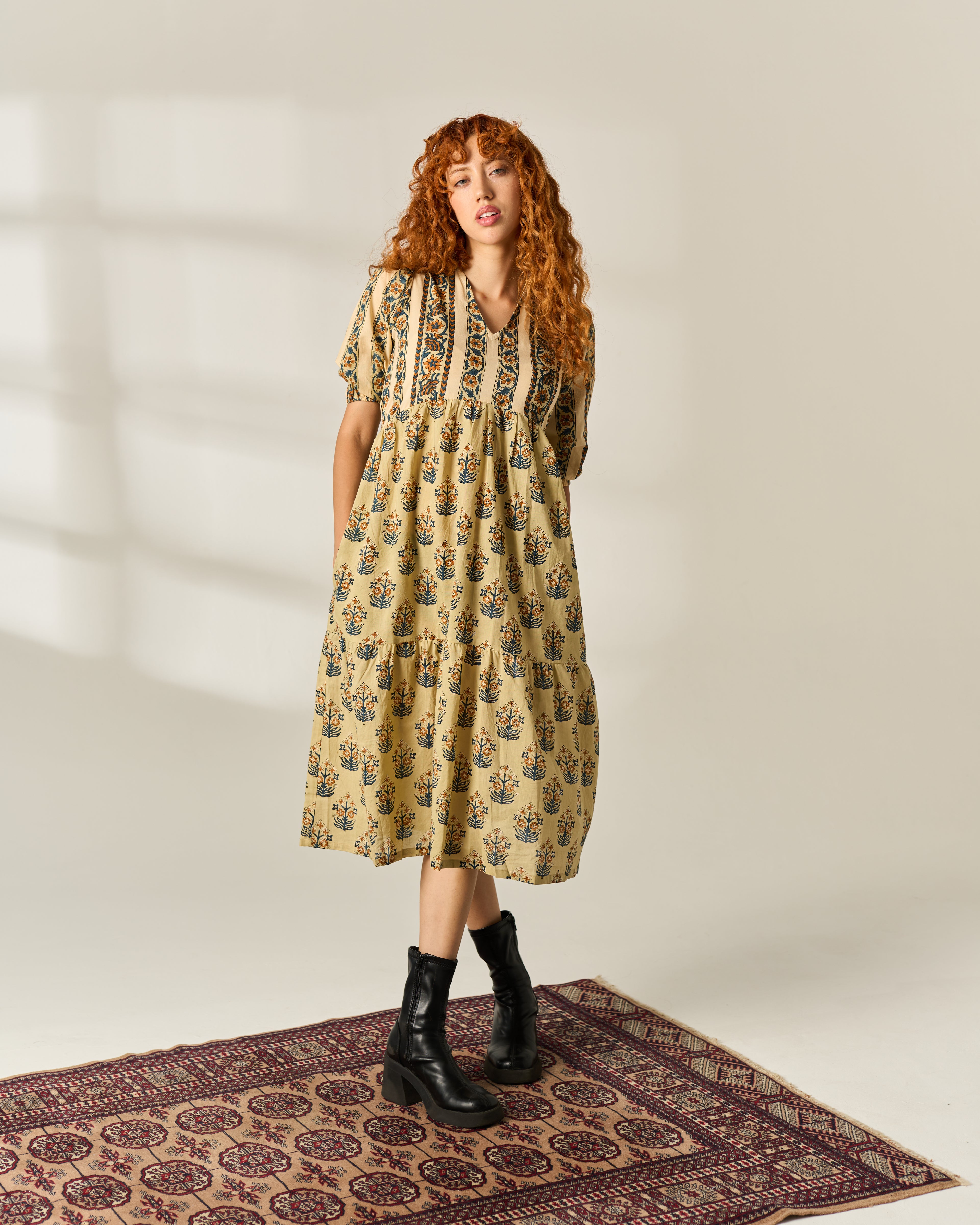 A woman with long curly red hair stands on a patterned rug wearing a beige midi dress with a blue and green floral hand block print, short puff sleeves, and black ankle boots against a warm off-white studio background.