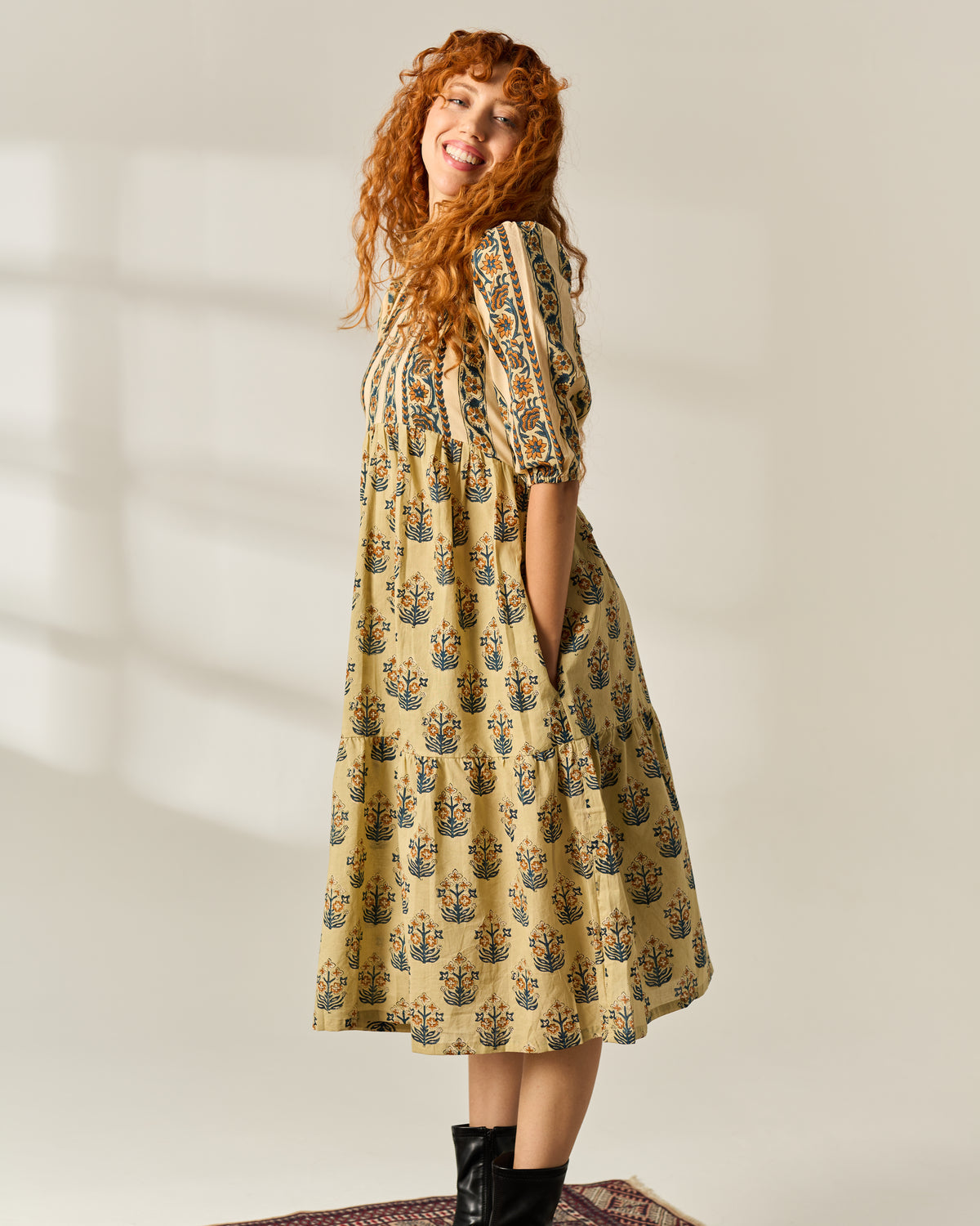 A woman with long curly red hair models a beige midi dress with a blue and green floral hand block print pattern. She is standing in a studio with soft window shadow lighting on a light beige background.