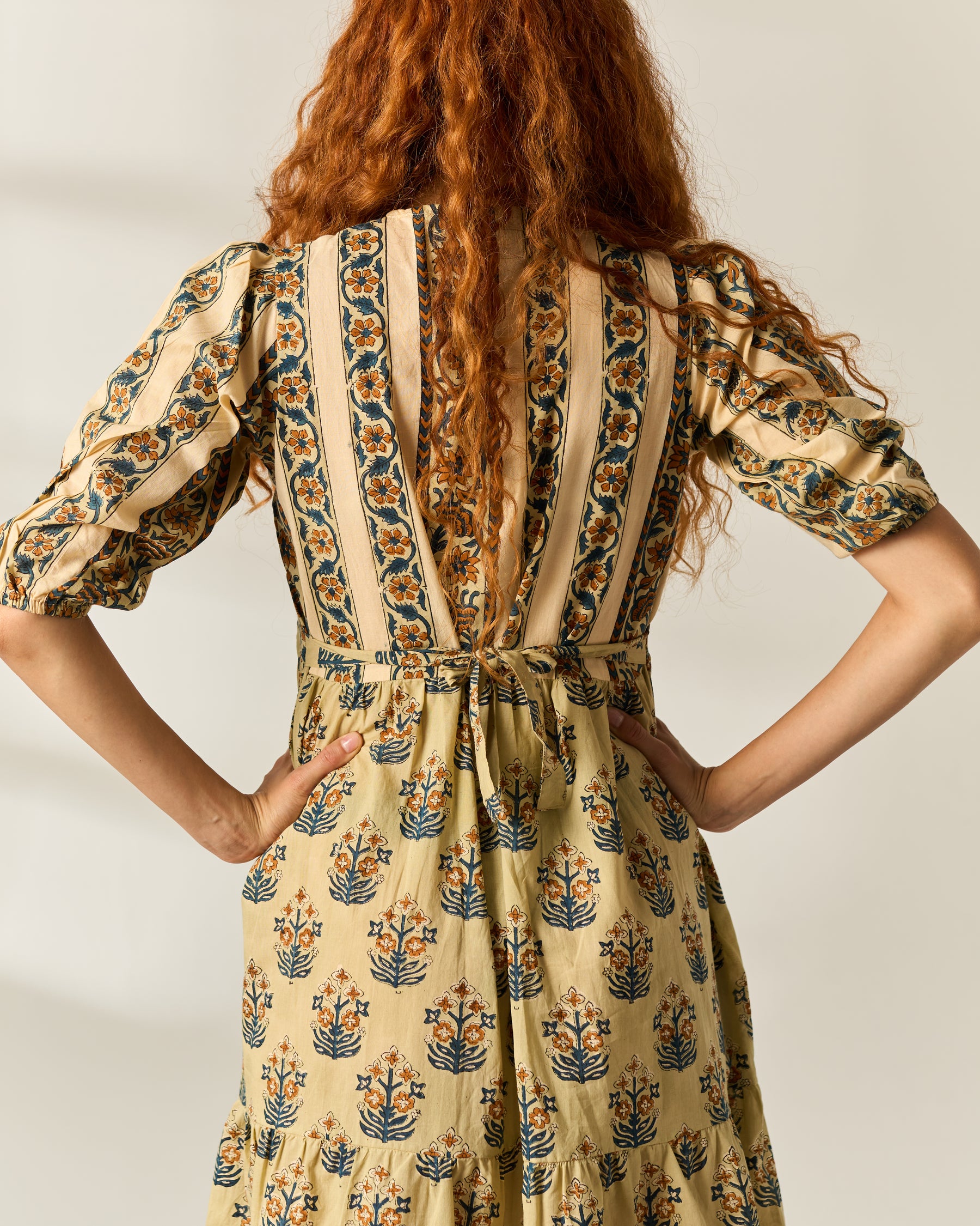 A woman viewed from behind wearing a cream-colored dress with vertical stripes and blue floral hand block prints, featuring puff sleeves and a tie waist.