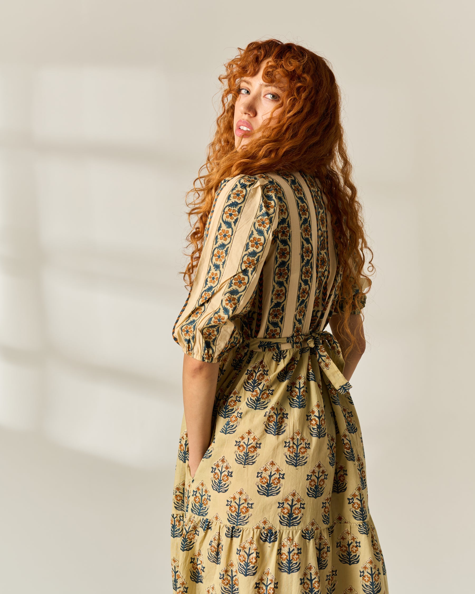 A woman with long, curly red hair is shown from the back wearing a beige tiered dress with a blue and orange floral hand block print pattern. She is photographed in a bright, minimalist studio setting with soft window shadows on the wall.