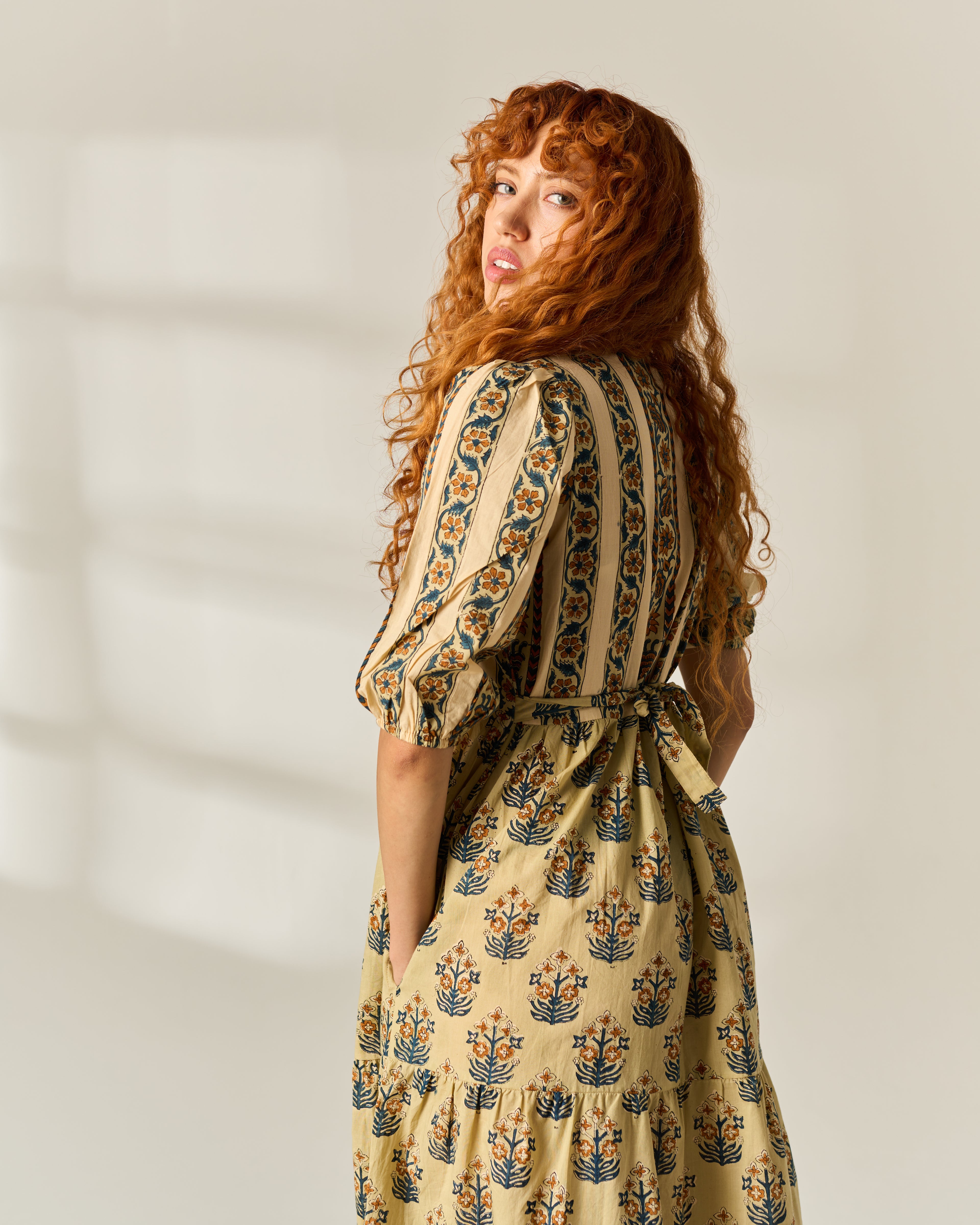 A woman with long, curly red hair is shown from the back wearing a beige tiered dress with a blue and orange floral hand block print pattern. She is photographed in a bright, minimalist studio setting with soft window shadows on the wall.
