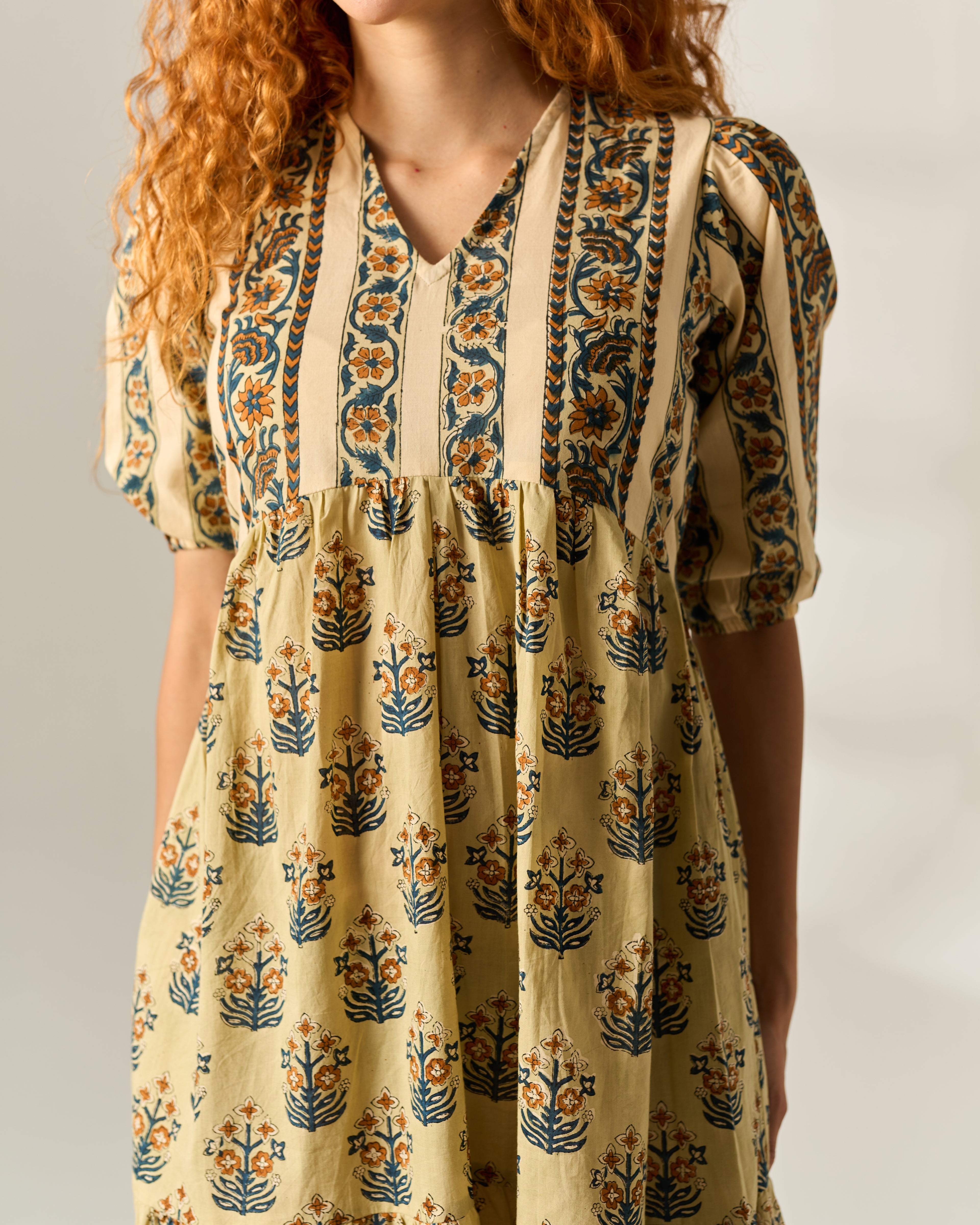 A woman wearing a short-sleeve V-neck dress with a beige base and blue, orange, and green floral hand block print, shown from the shoulders to the hips against a plain light background.
