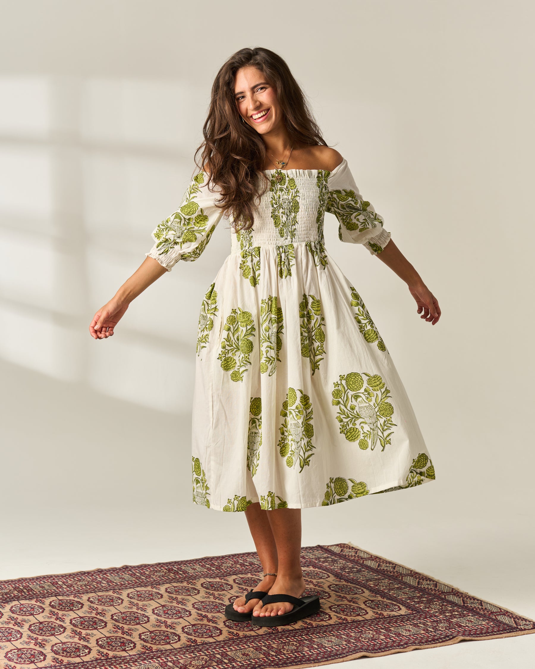 A woman modeling an off-the-shoulder midi dress with a green floral hand block print on an off-white 100% cotton fabric, standing on a patterned rug against a natural background.
