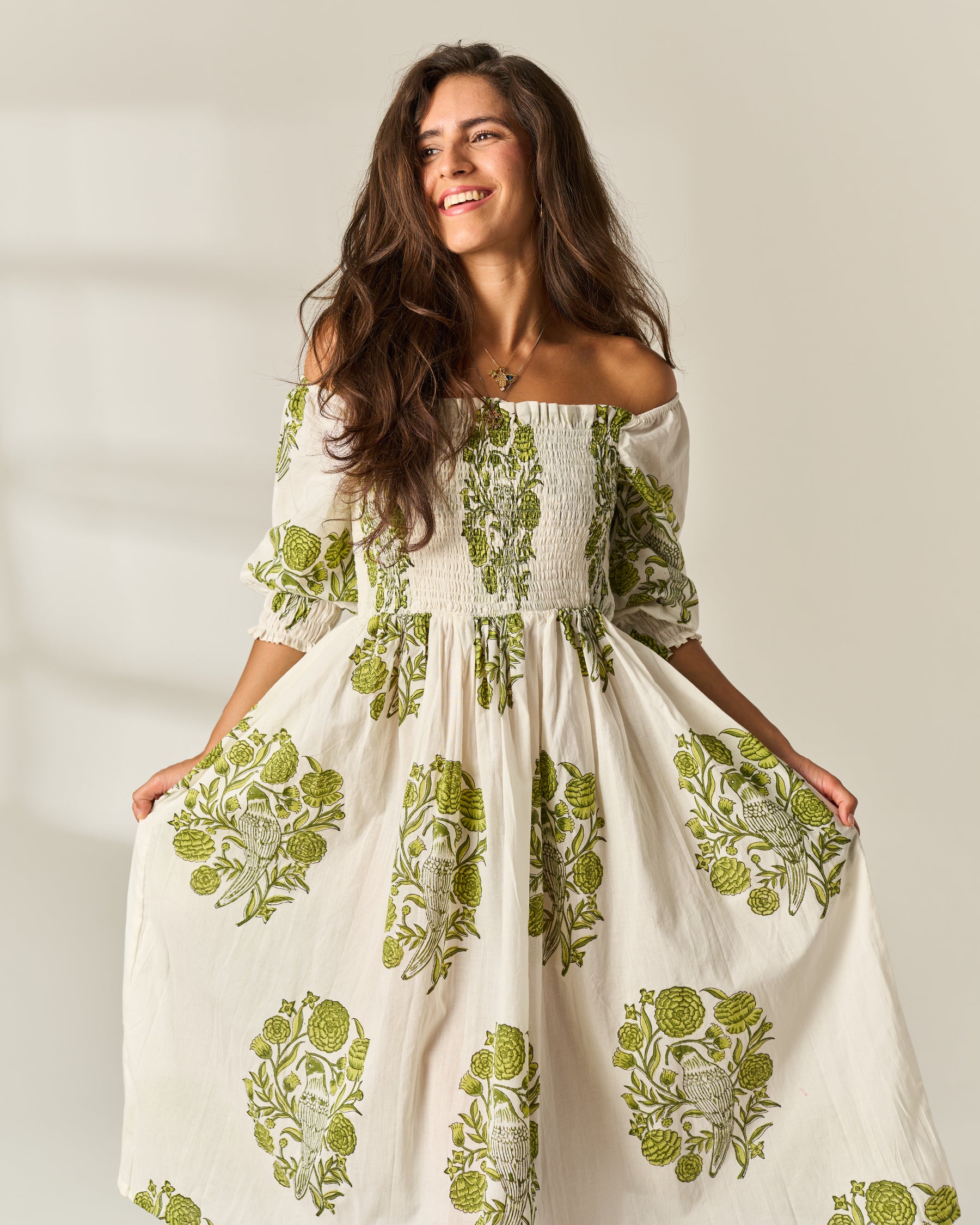 A woman wearing a cream off-the-shoulder hand block painted cotton maxi dress with green botanical block-print patterns, smiling and holding out the skirt against a warm, softly lit beige background.