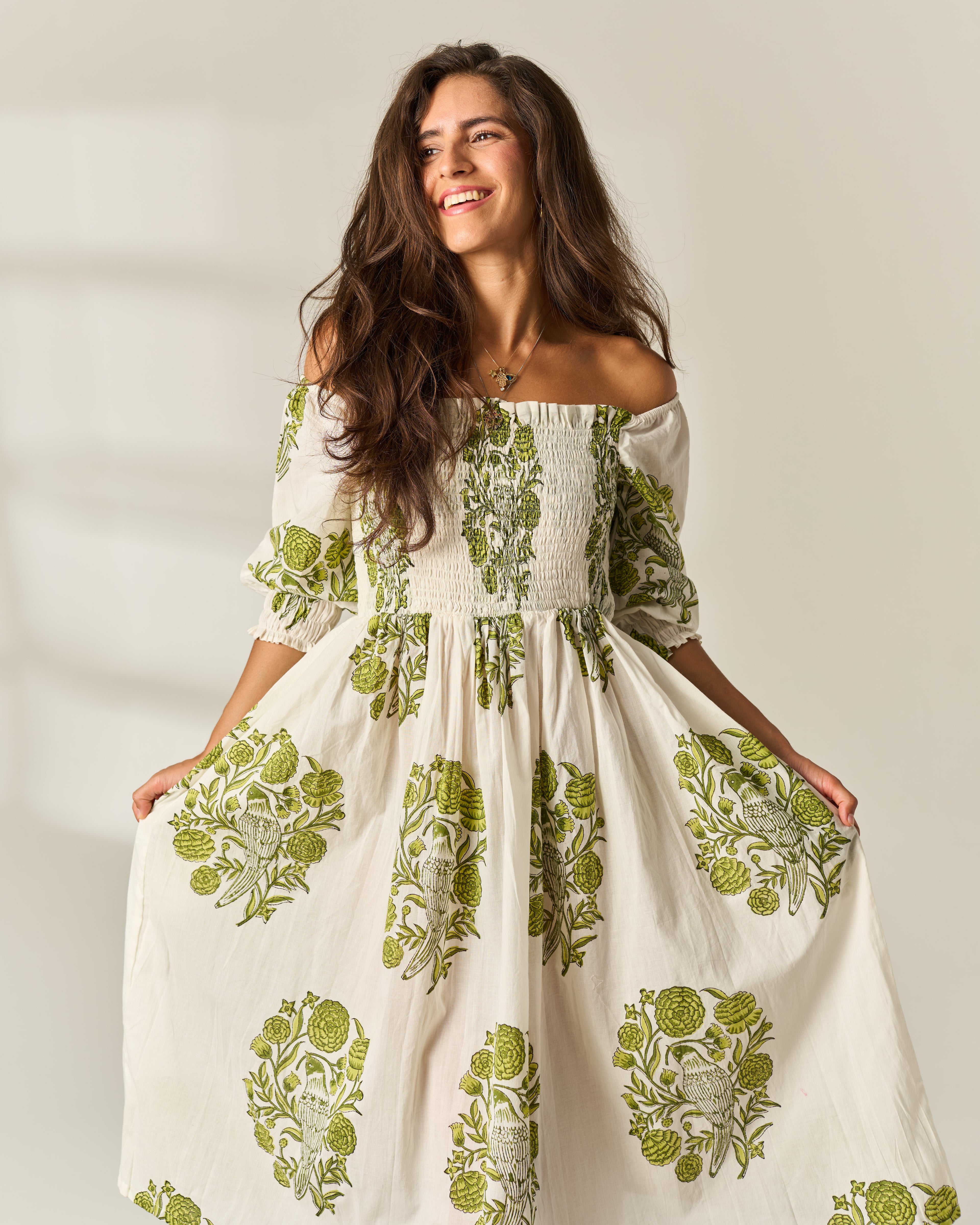 A woman wearing a cream off-the-shoulder hand block painted cotton maxi dress with green botanical block-print patterns, smiling and holding out the skirt against a warm, softly lit beige background.