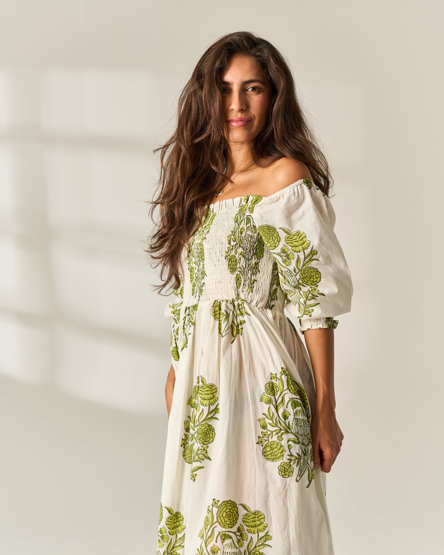 A woman wearing an off-the-shoulder midi dress with a white base and green botanical floral hand block print, standing against a warm off-white background with soft window shadows.