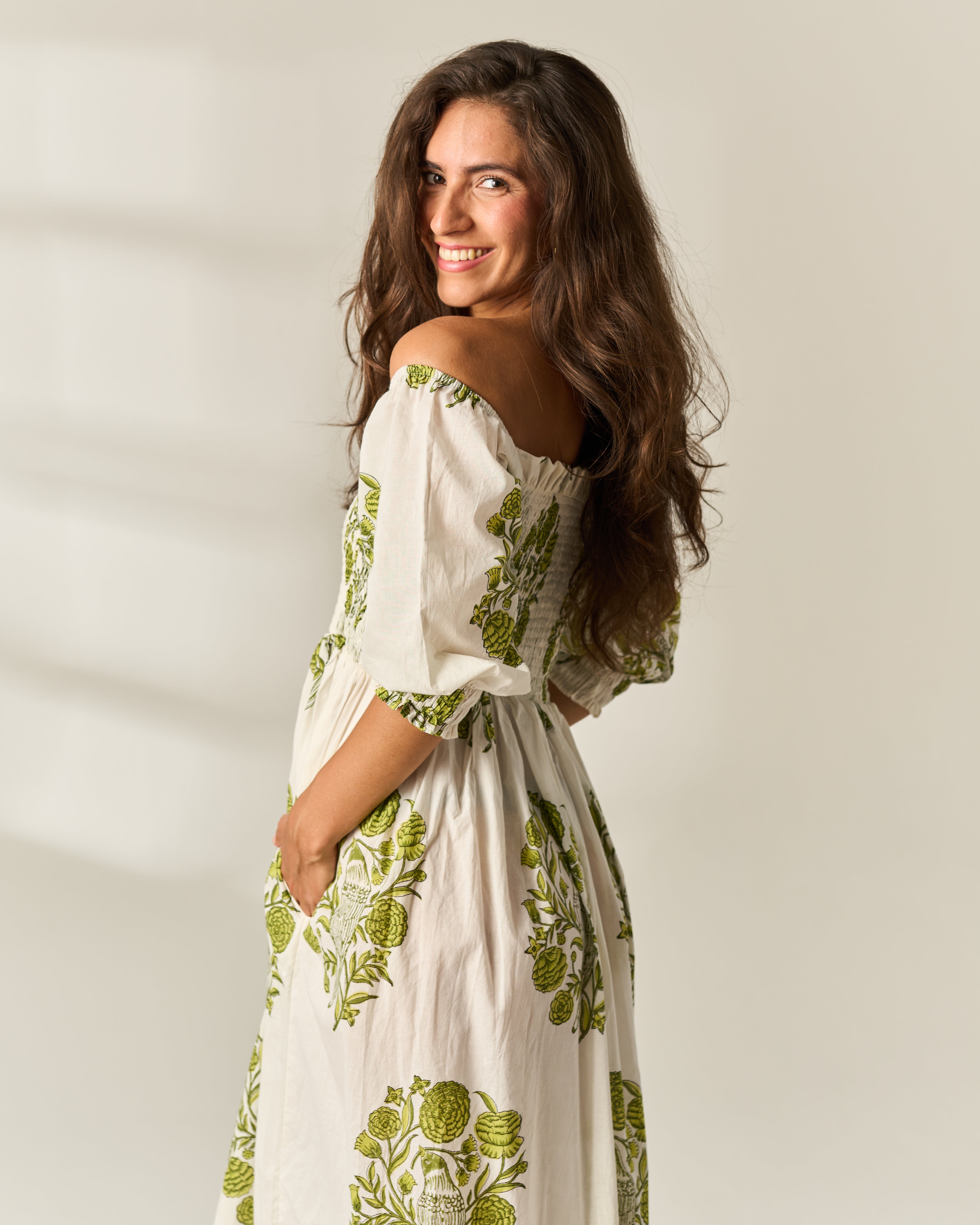 A woman wearing an off-the-shoulder midi dress with a green botanical hand block print on a cream-colored 100% cotton fabric, posing against a warm off-white background.