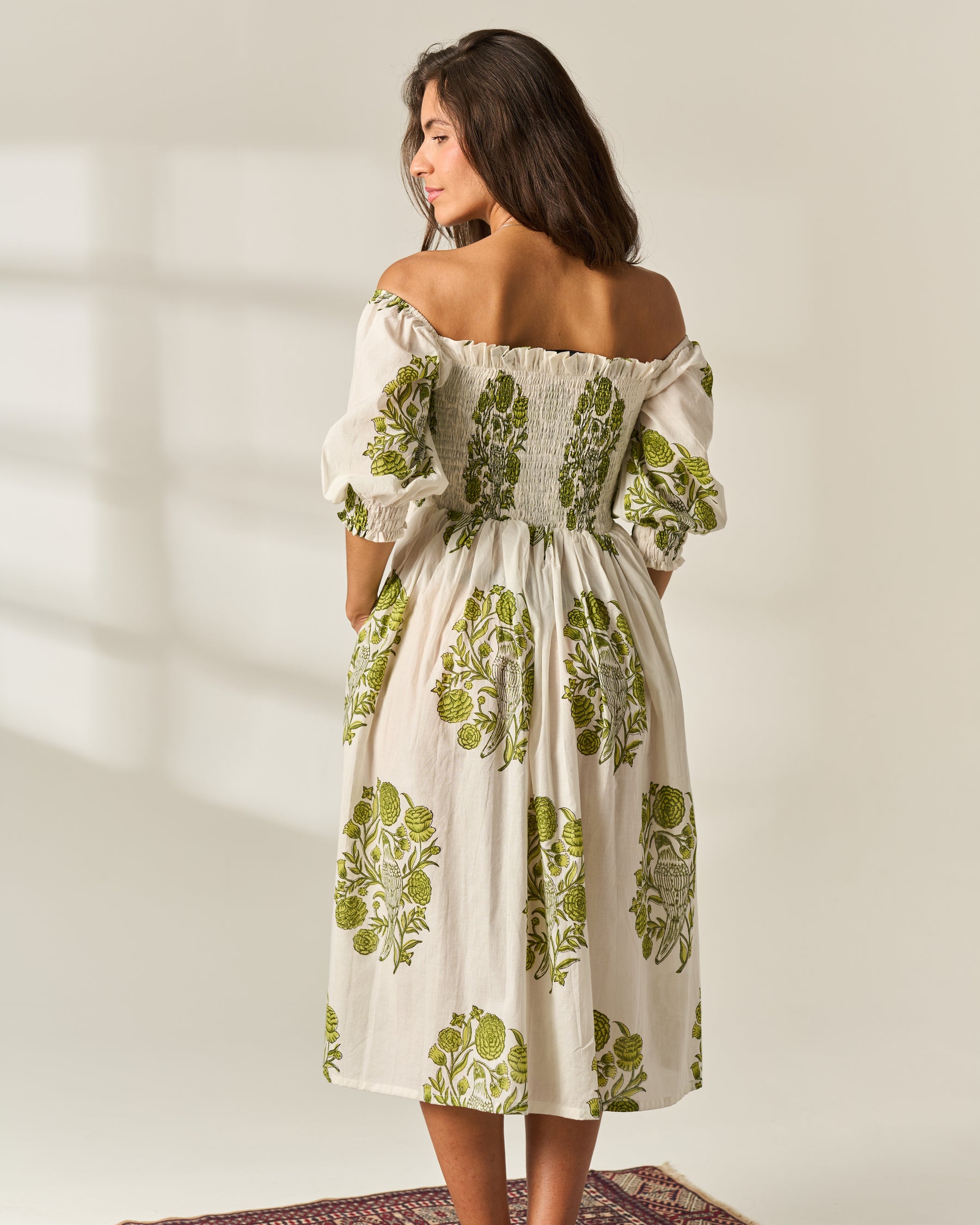 A woman wearing an off-the-shoulder midi dress with a green botanical Hand Block print on a cream-colored fabric, shown from the back against a warm beige background.