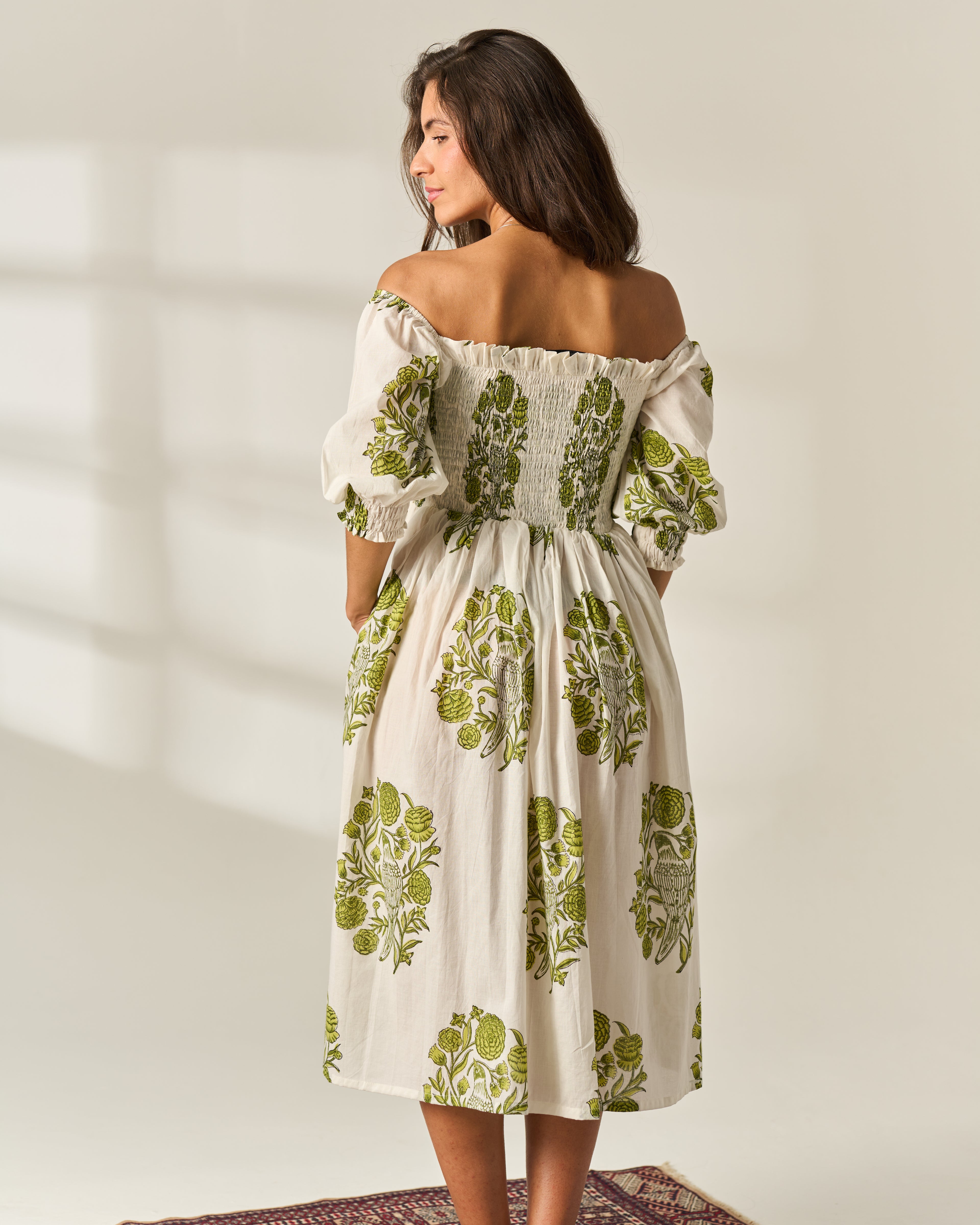 A woman wearing an off-the-shoulder midi dress with a green botanical Hand Block print on a cream-colored fabric, shown from the back against a warm beige background.