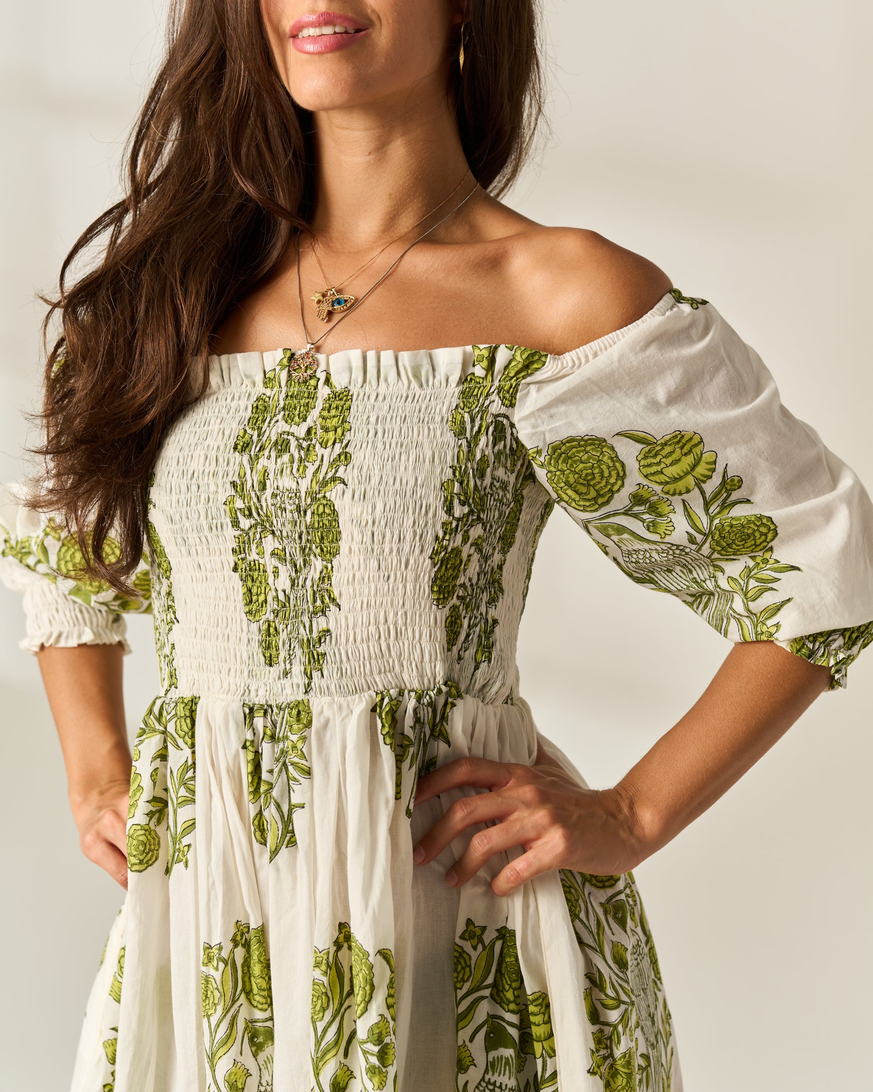 A woman wearing an off-the-shoulder white dress with green botanical floral Hand Block prints, featuring a smocked bodice and puff sleeves.