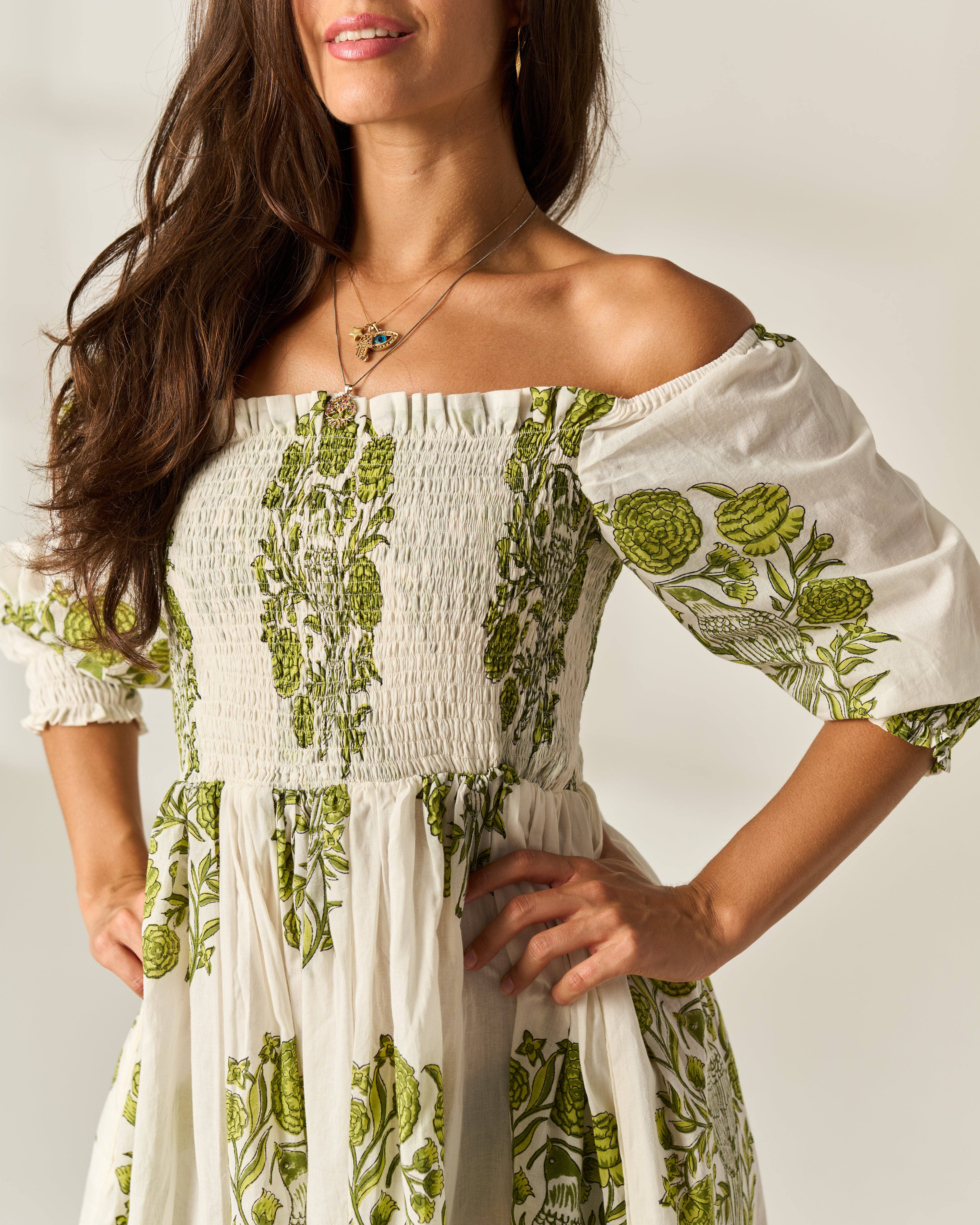 A woman wearing an off-the-shoulder white dress with green botanical floral Hand Block prints, featuring a smocked bodice and puff sleeves.