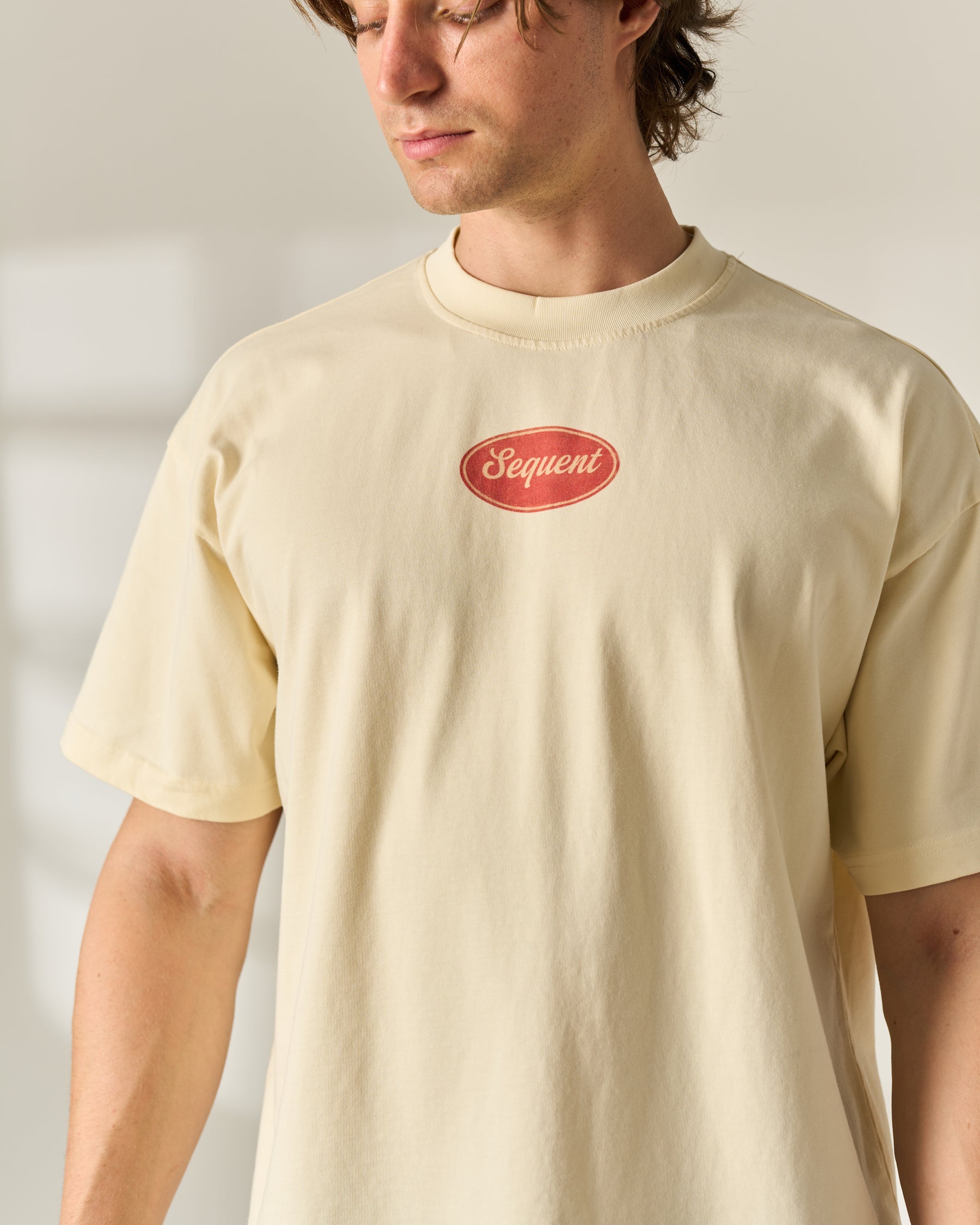 A man wearing a cream-colored crewneck t-shirt with a red oval 'Sequent' logo on the chest, photographed in a bright, minimalist indoor setting.
