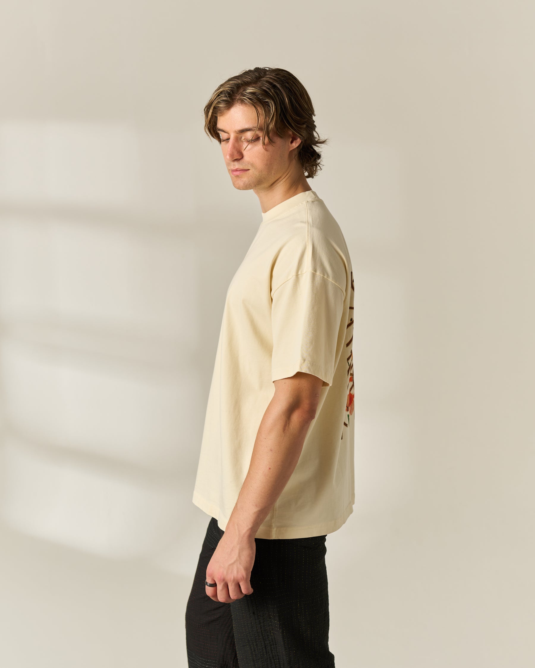 A man wearing a cream-colored short-sleeve t-shirt with a graphic print on the back, paired with black pants, standing in profile against a warm beige background.