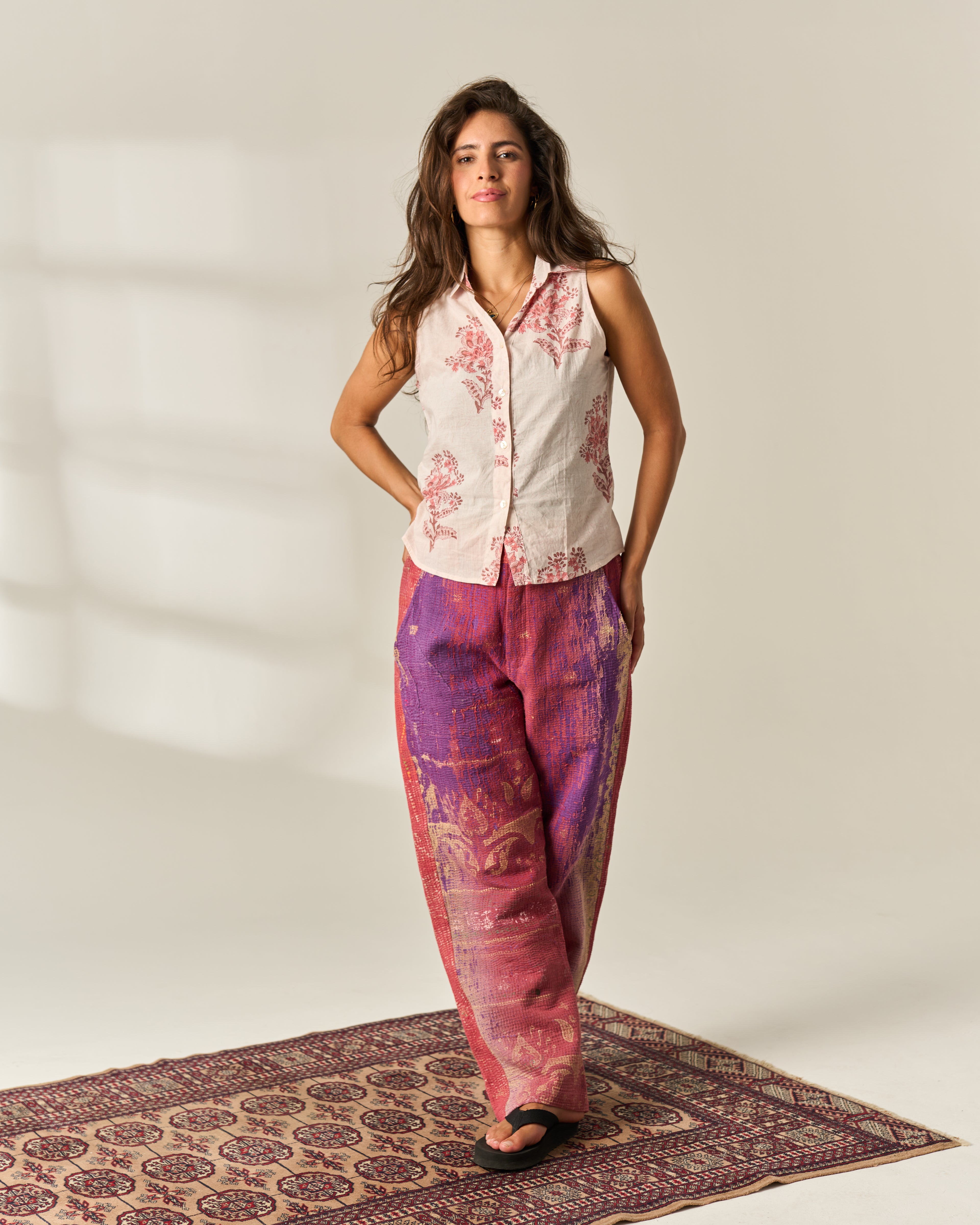 A woman stands on a patterned rug in a studio wearing a sleeveless white button-up Hand Block Paintedshirt with pink floral print and loose purple-pink printed vintage upcycled kantha pants.