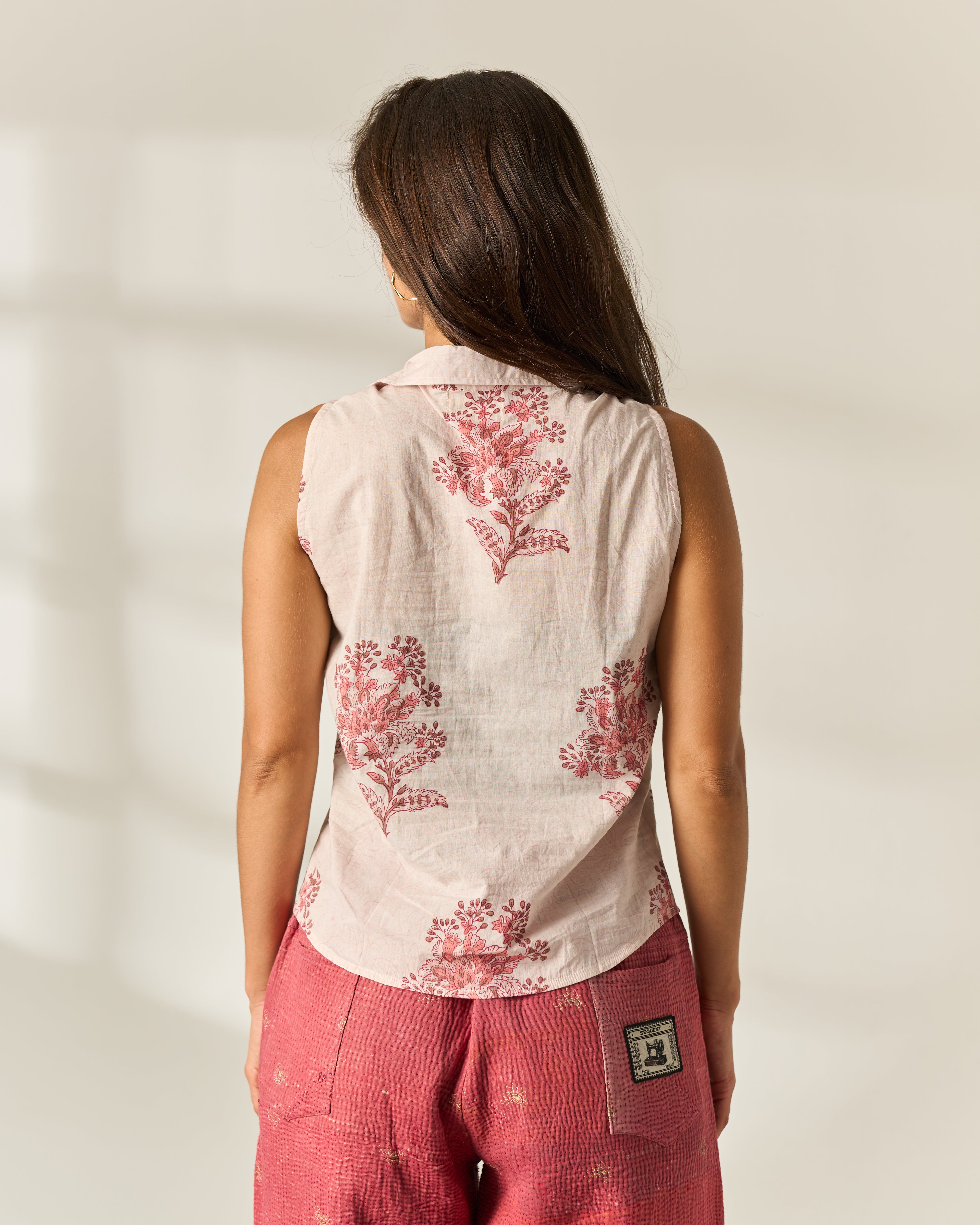 A woman viewed from behind wearing a sleeveless light pink 100% cotton, hand block Painted Shirt with a red floral print and red textured upcycled Vintage Kantha pants, standing against a warm off-white background.