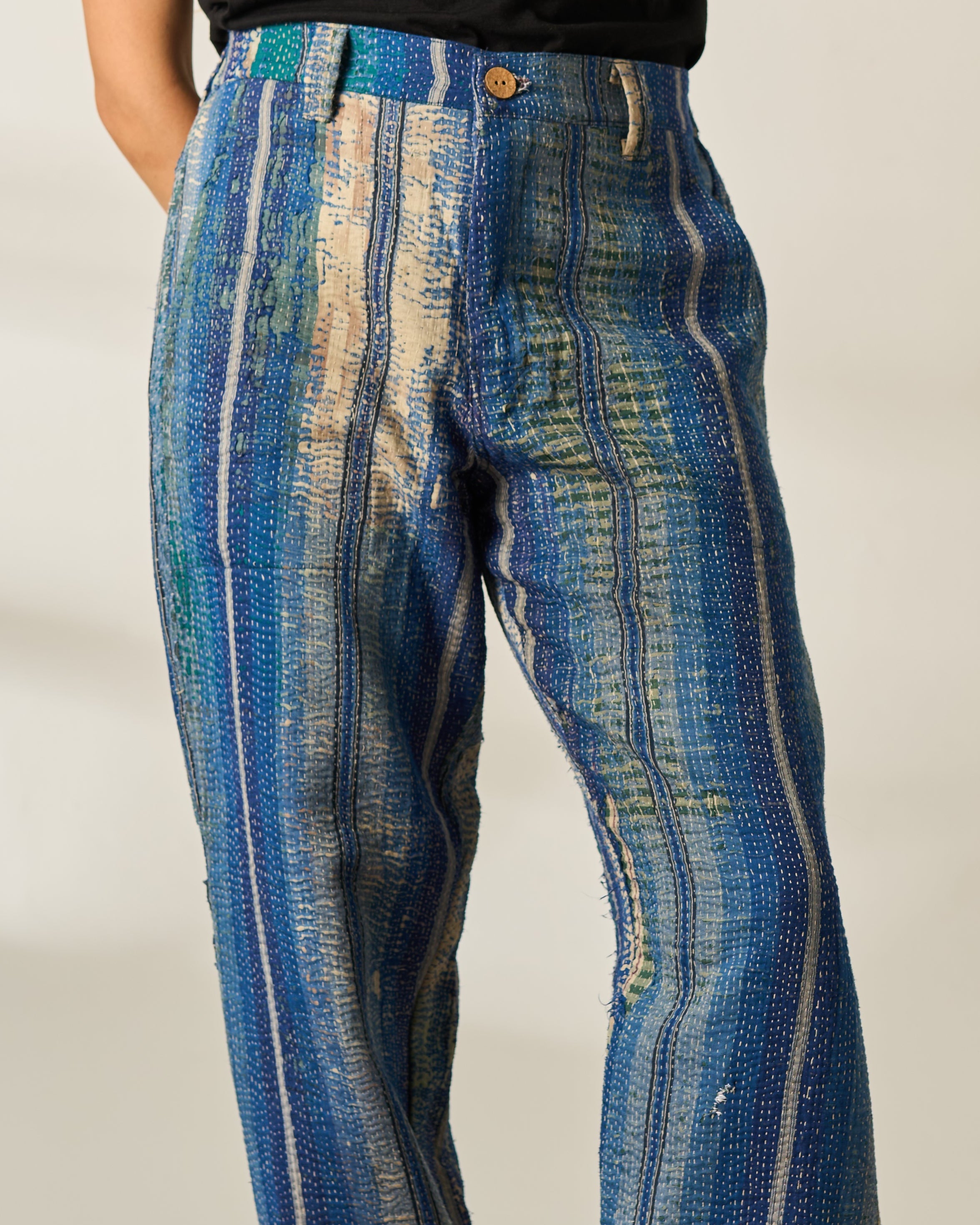 A person wearing blue patterned wide leg vintage kantha trousers with vertical textured stripes in blue, beige, and gold, paired with a black top and black leather boots, standing on a patterned rug against a plain beige wall.