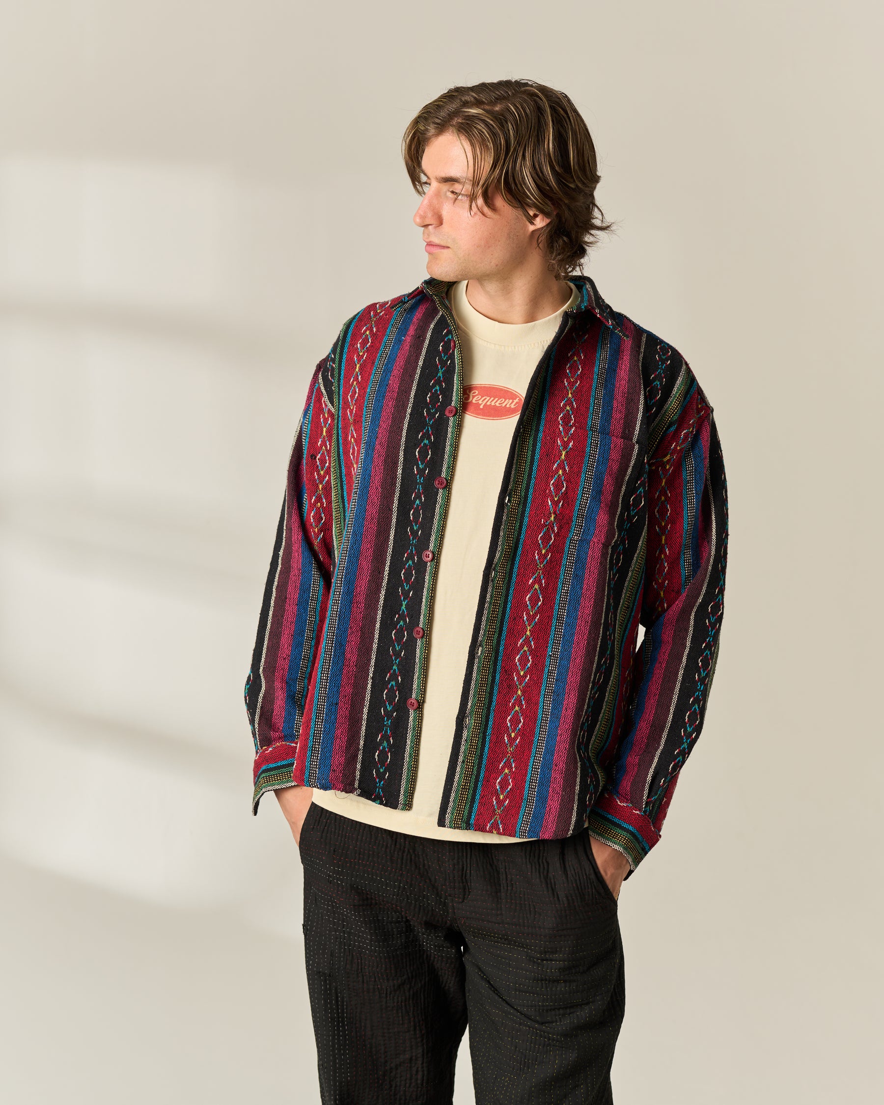 A man wearing a colorful striped button-up Jacquard Over-shirt over a cream t-shirt and dark pants, standing against a neutral beige background.