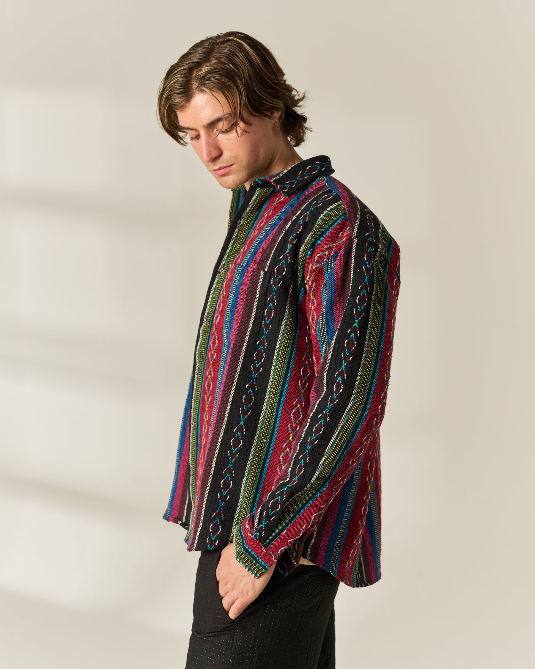 A man wearing a long-sleeve button-up Jacquard Over-shirt with a vibrant multicolor vertical stripe pattern, paired with black trousers, photographed against a warm off-white background.