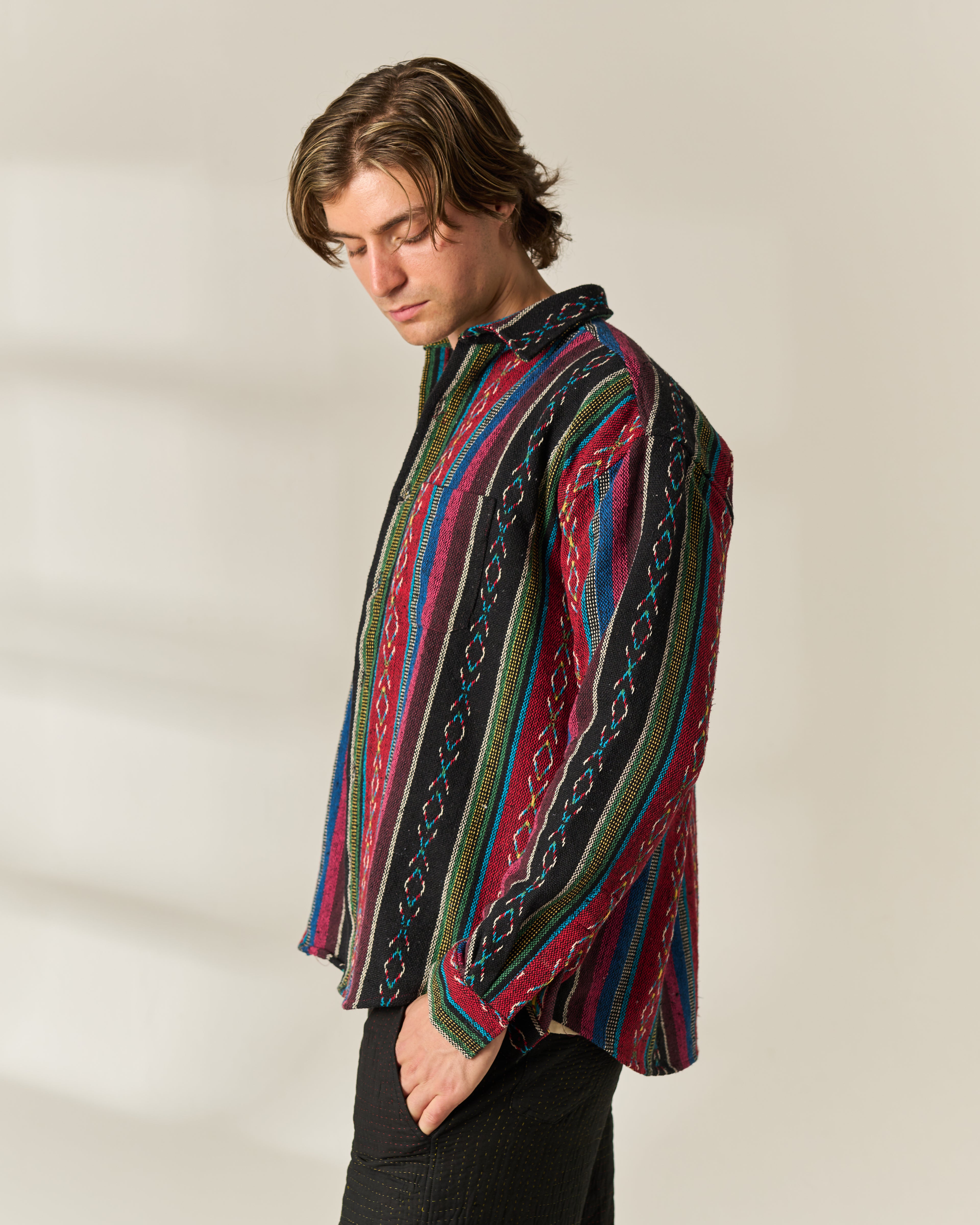 A man wearing a long-sleeve button-up Jacquard Over-shirt with a vibrant multicolor vertical stripe pattern, paired with black trousers, photographed against a warm off-white background.