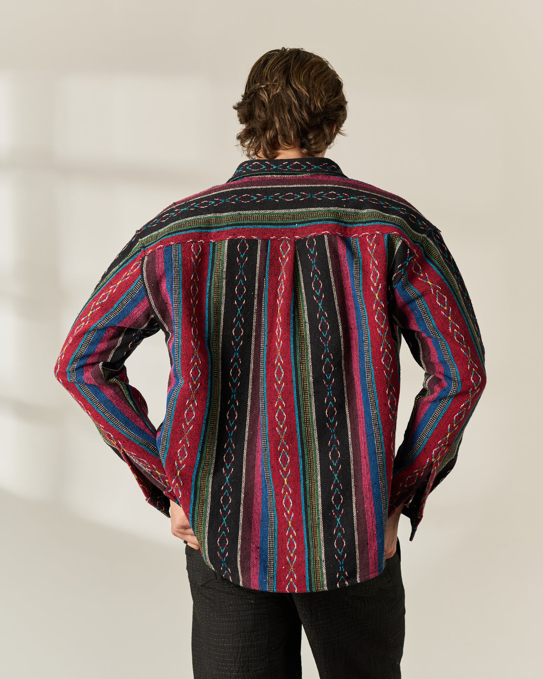 A man viewed from behind wearing a long-sleeve button-up Jacquard Over-shirt with a vibrant multicolor vertical stripe pattern in red, blue, green, and black, paired with black pants.