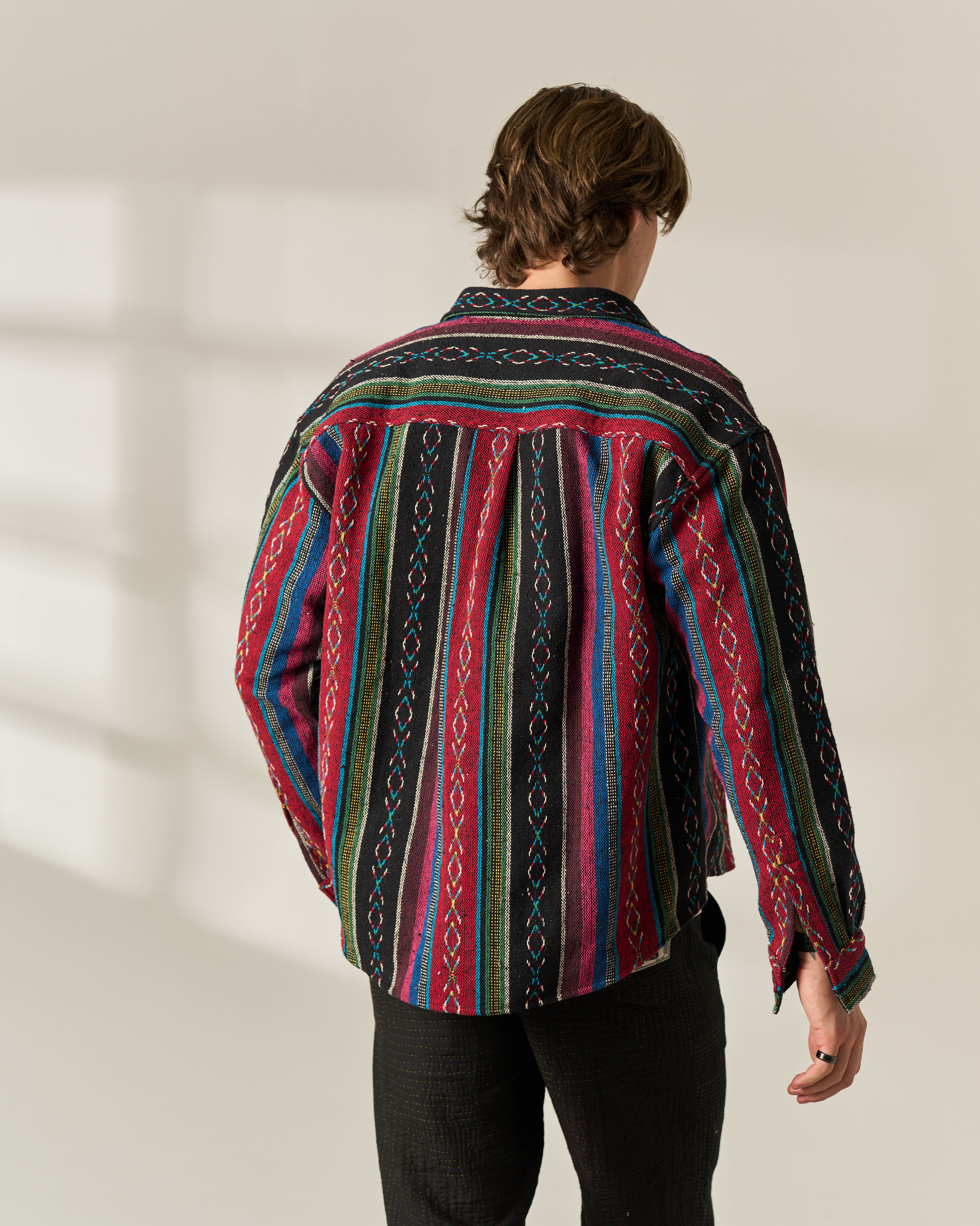 A man viewed from behind wearing a long-sleeve button-up Jacquard Over-shirt with a vibrant multicolor geometric pattern in red, blue, green, and black, paired with black pants against a warm off-white background.