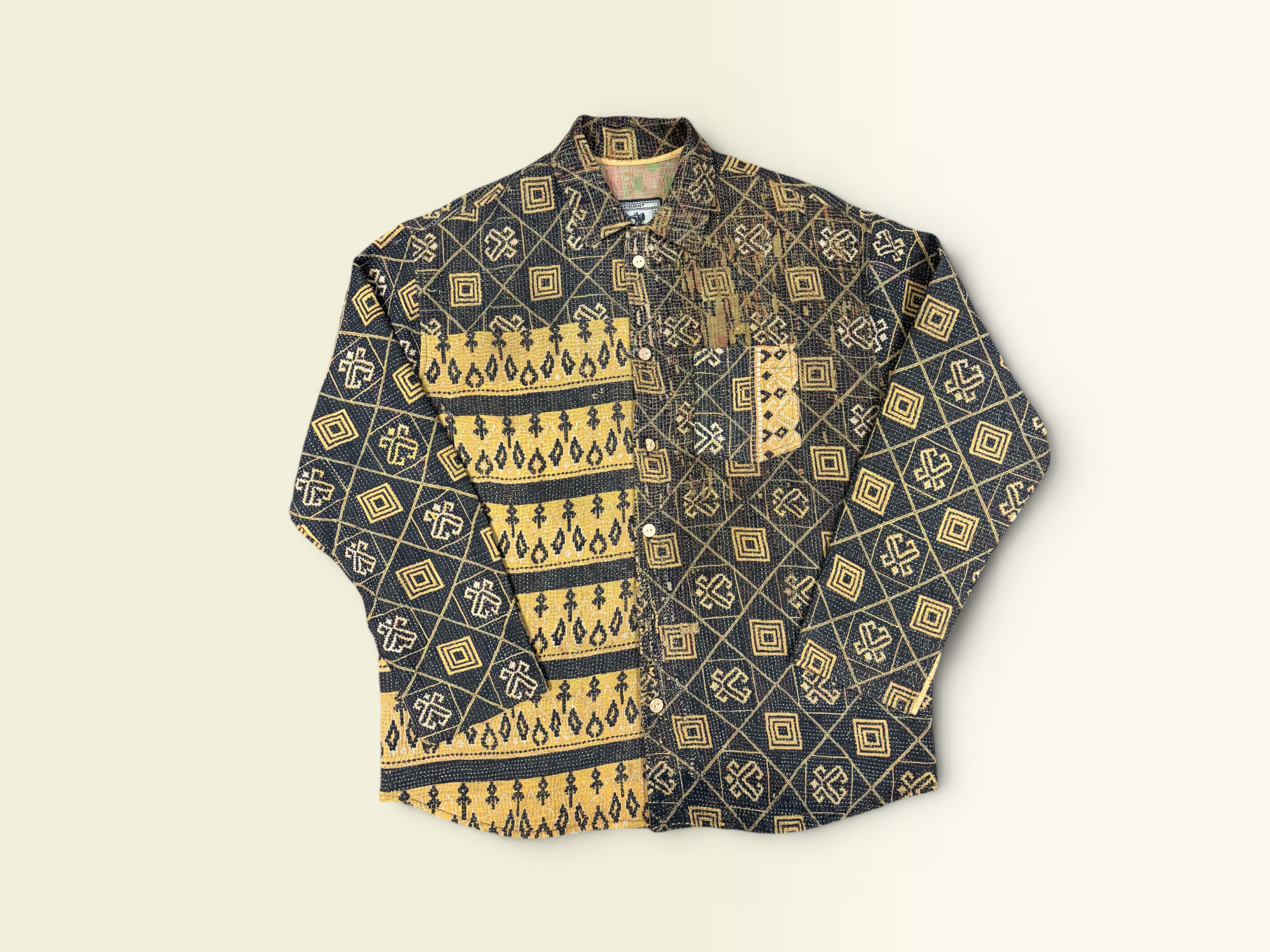 A long-sleeve button-up vintage Kantha shirt with a complex geometric and tribal-inspired pattern in mustard yellow, beige, and black, laid flat against a light cream background.