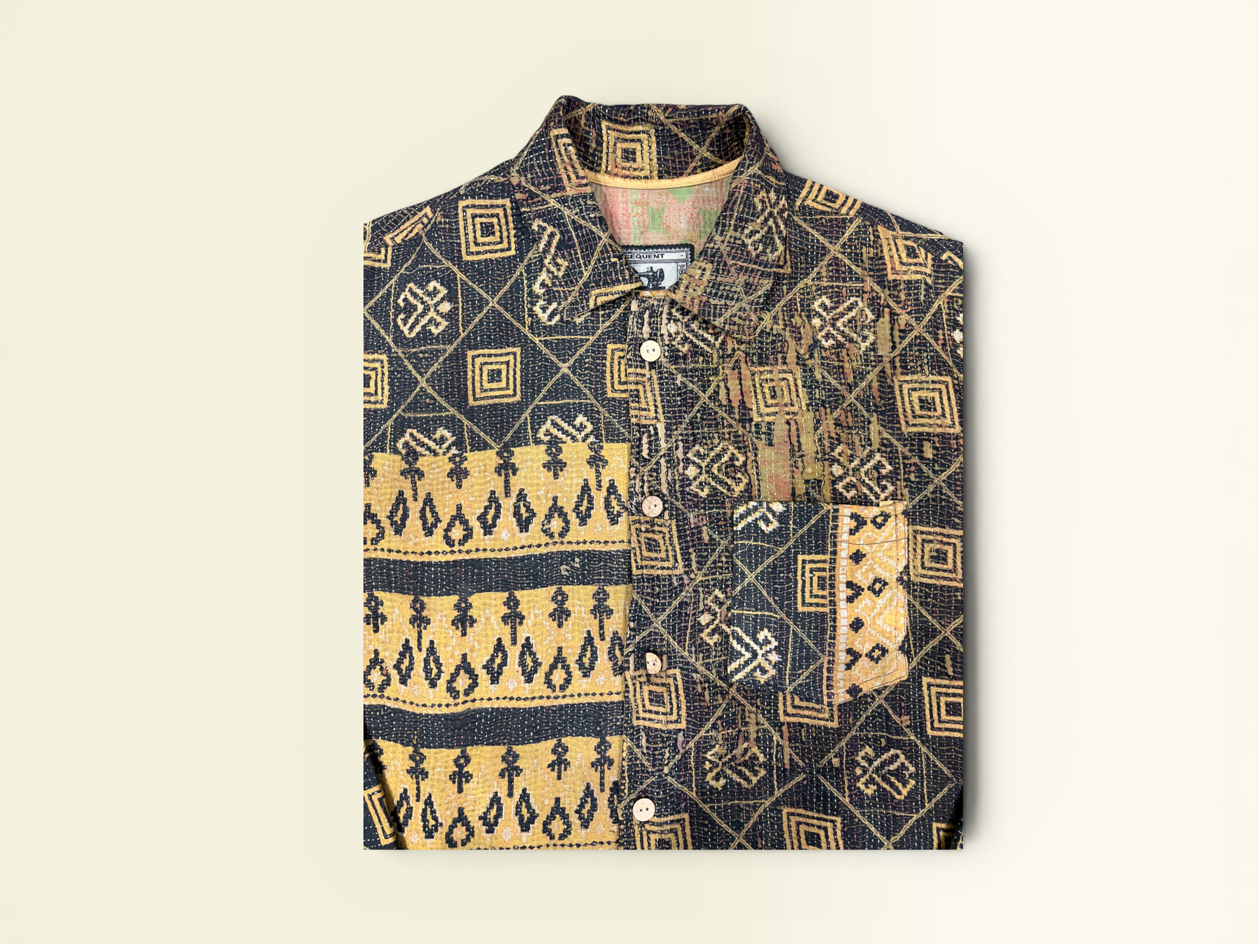 A folded button-up vintage Kantha shirt with a dark navy and tan patchwork pattern featuring geometric and tribal-inspired motifs, displayed on a light beige background.