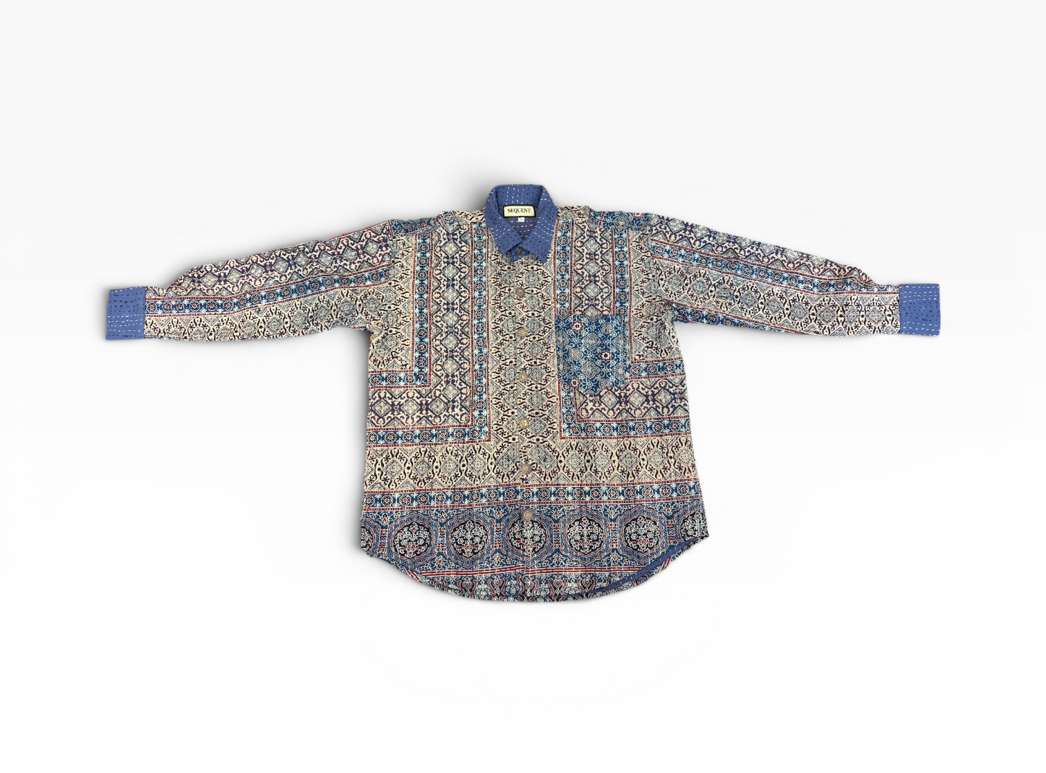A long-sleeve button-up Ajrakh Kantha hand painted and hand stitched shirt with an intricate geometric pattern in shades of indigo, beige, and brown, laid flat against a solid white background. Blue cuffs and blue collar. 220 GSM Cotton fabric construction.