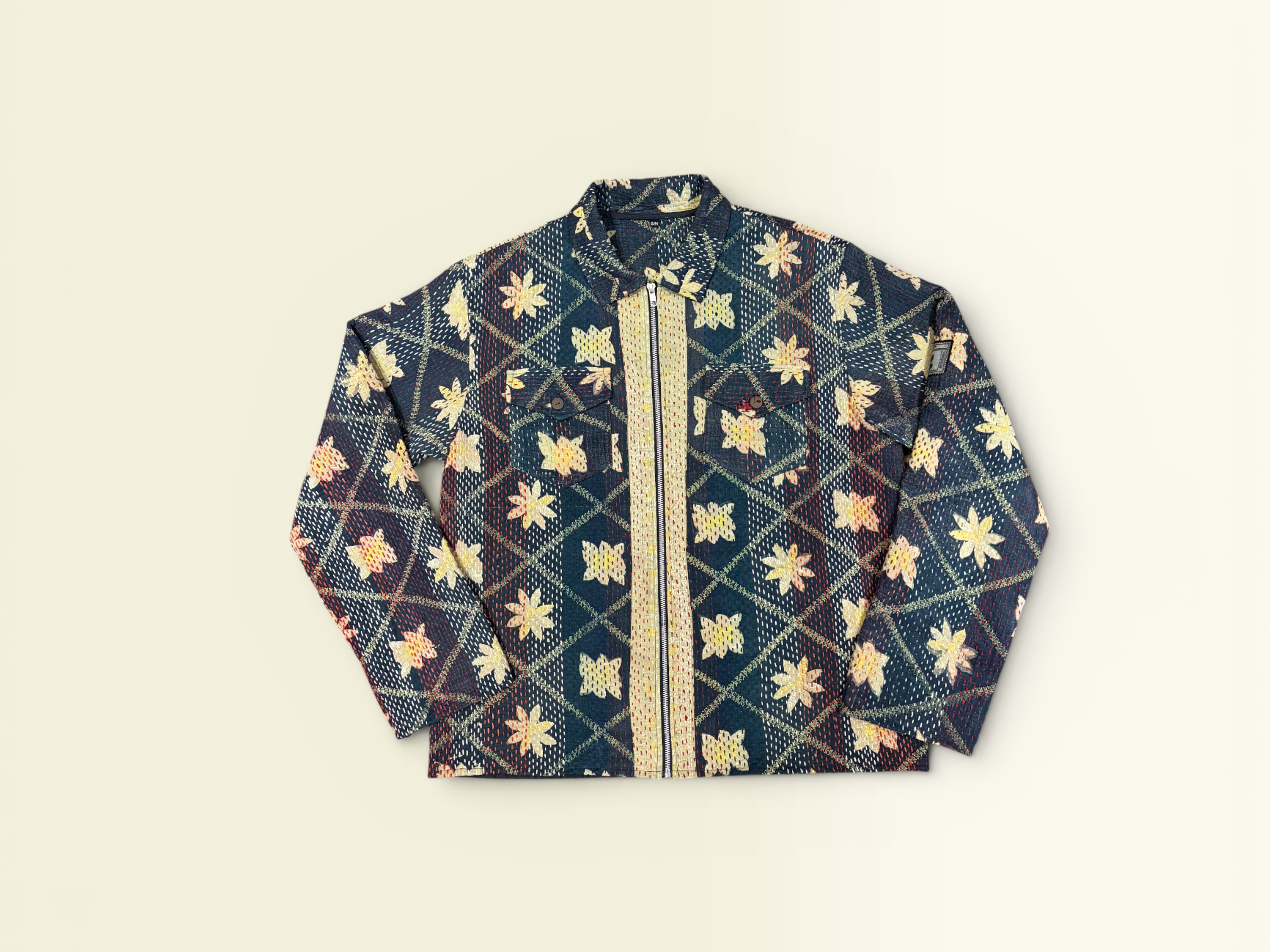 A Vintage Kantha long-sleeve jacket with a dark navy blue base, decorated with a cream and gold floral pattern and a central YKK zipper, laid flat against a light beige background.