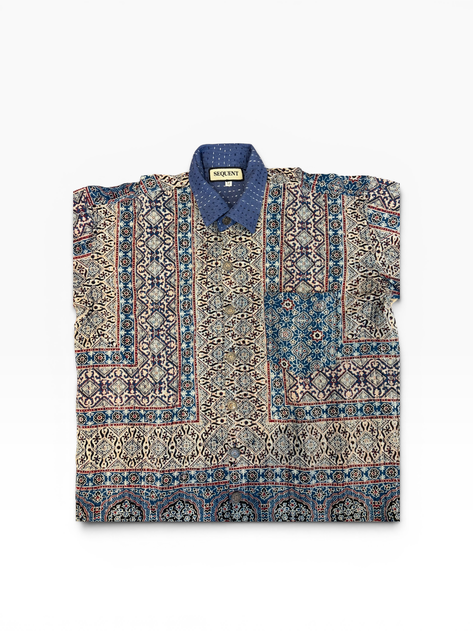 A folded short-sleeve button-up Ajrakh Kantha shirt with an intricate multi-color geometric and tribal pattern, featuring a solid blue collar and a visible 'Sequent' brand label.